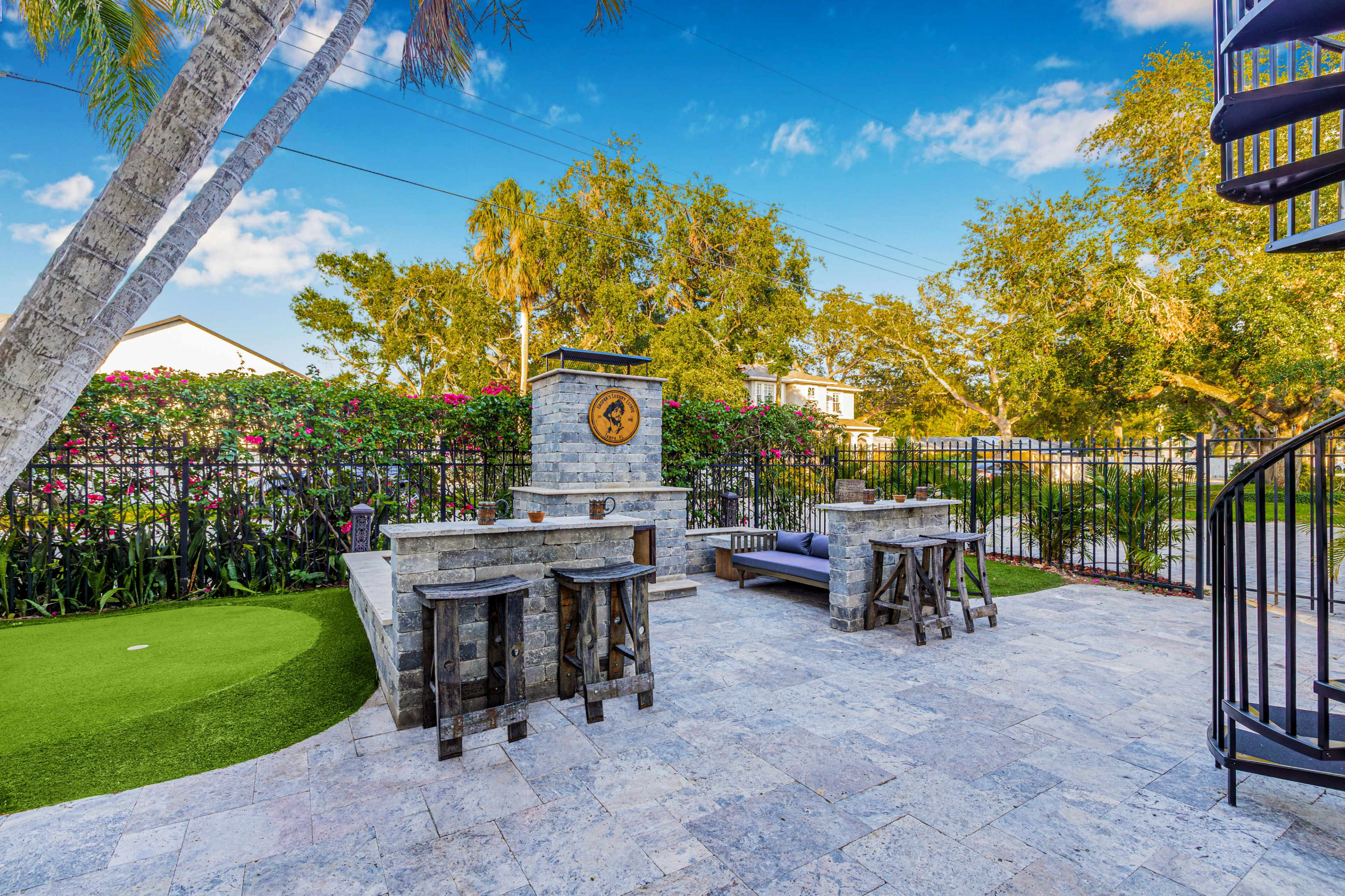 A stone patio features a built-in grill and seating surrounded by lush greenery and a spiral staircase in the background.