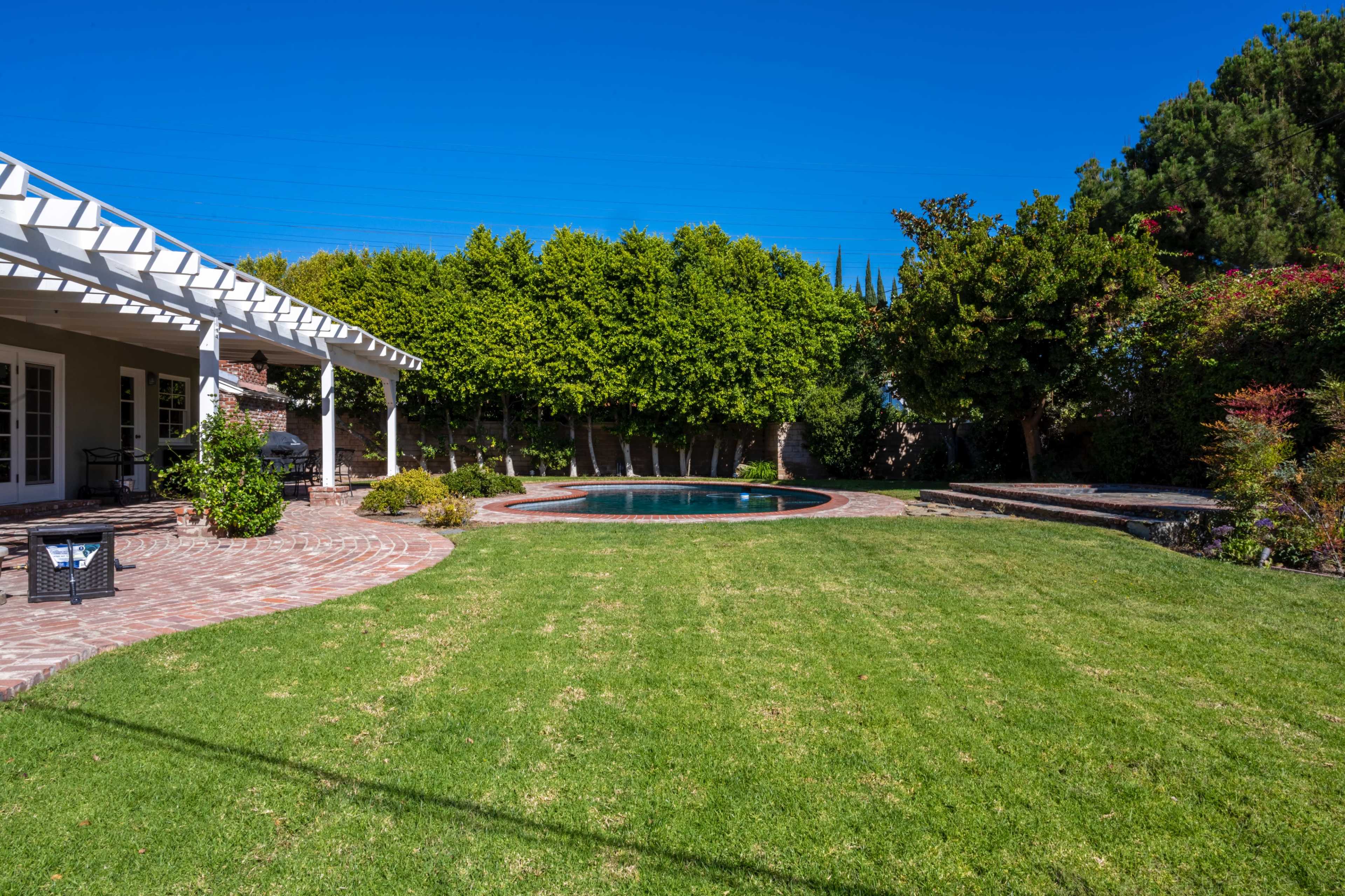 A backyard features a swimming pool surrounded by neatly trimmed grass and landscaping, with a covered patio and bright blue sky overhead.