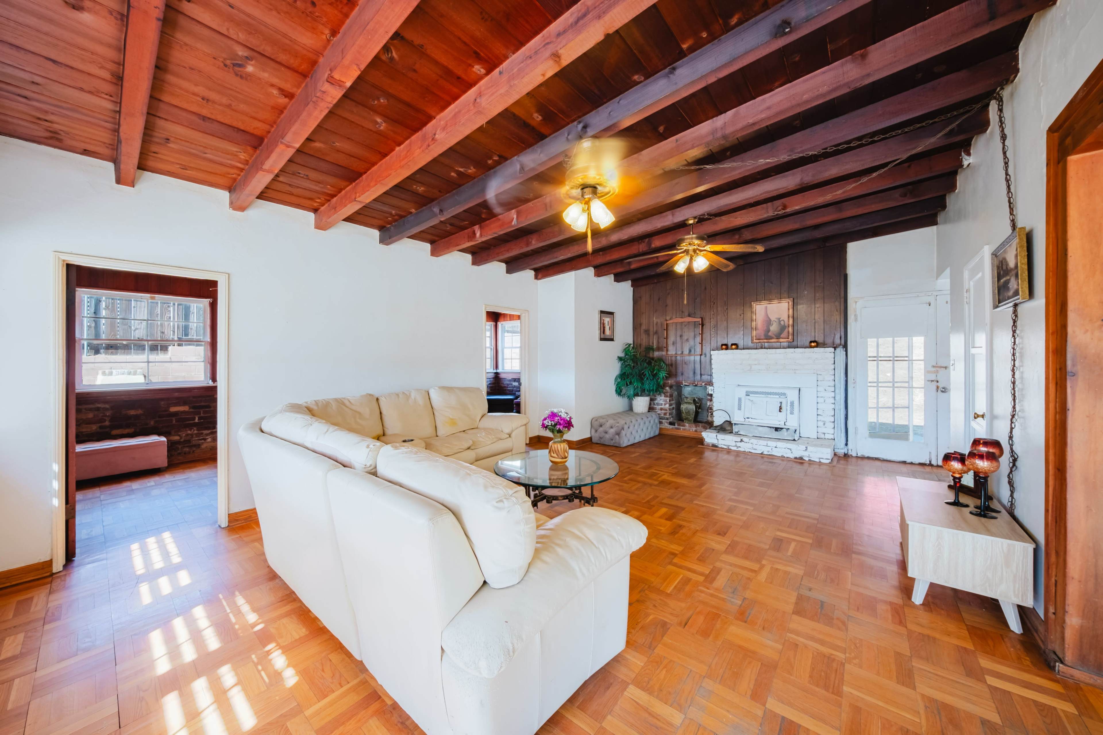 A spacious living room features a white leather sofa, wooden ceiling beams, and a fireplace against a dark wood accent wall.