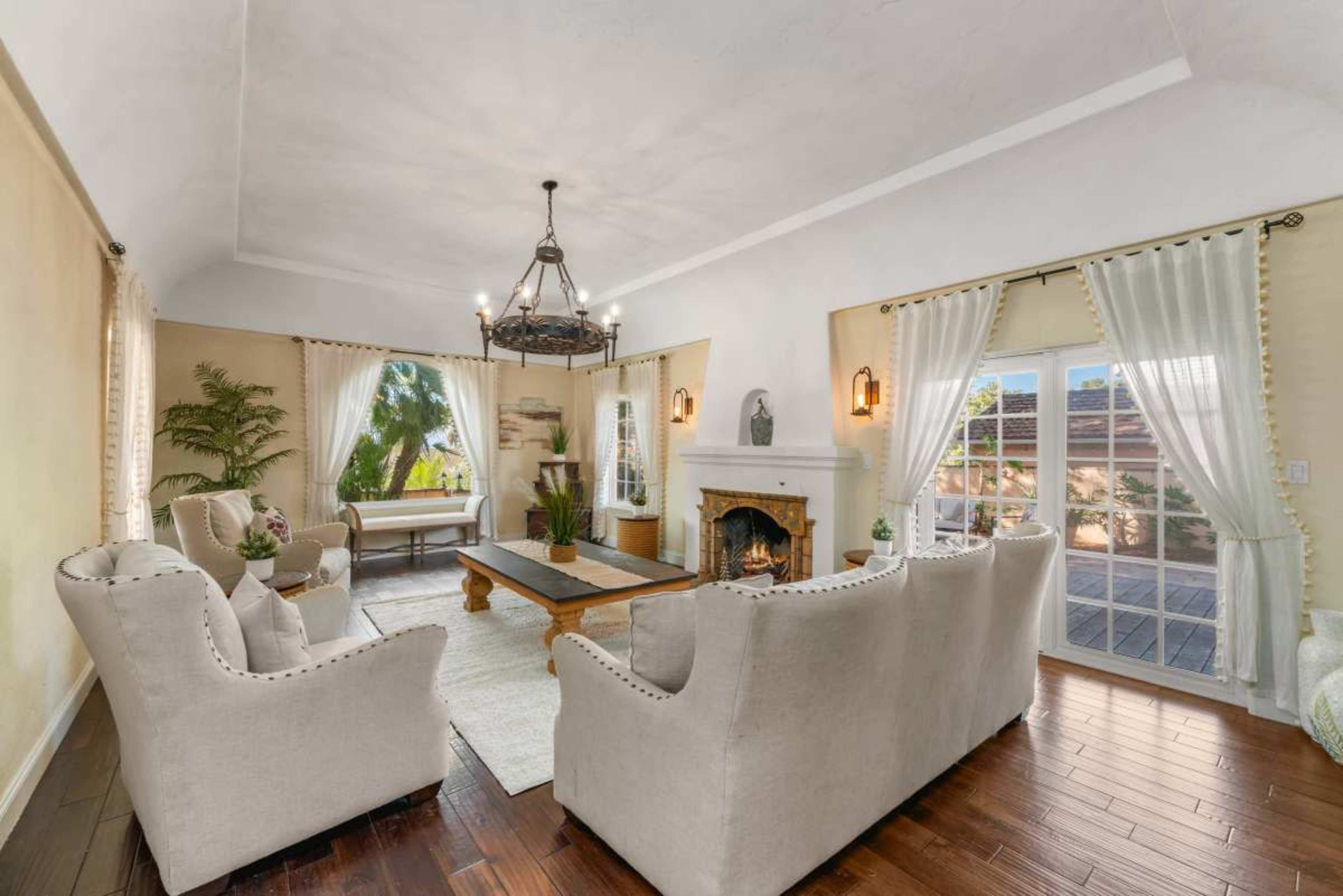 The image shows a spacious living room with two light-colored chairs, a wooden coffee table, a fireplace, and large windows that let in natural light.