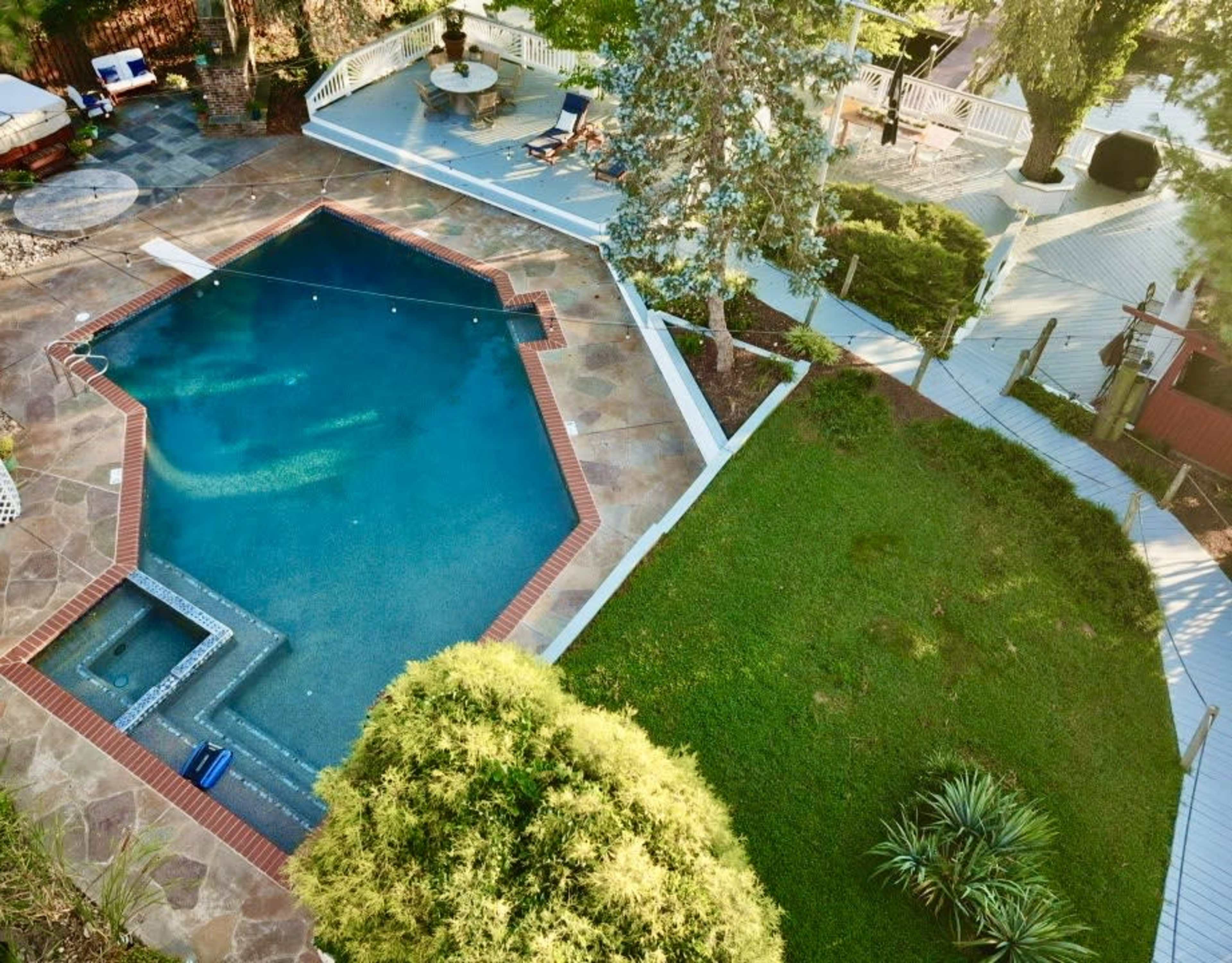 An aerial view shows a pool with a hot tub, surrounded by a stone patio and landscaped lawn.