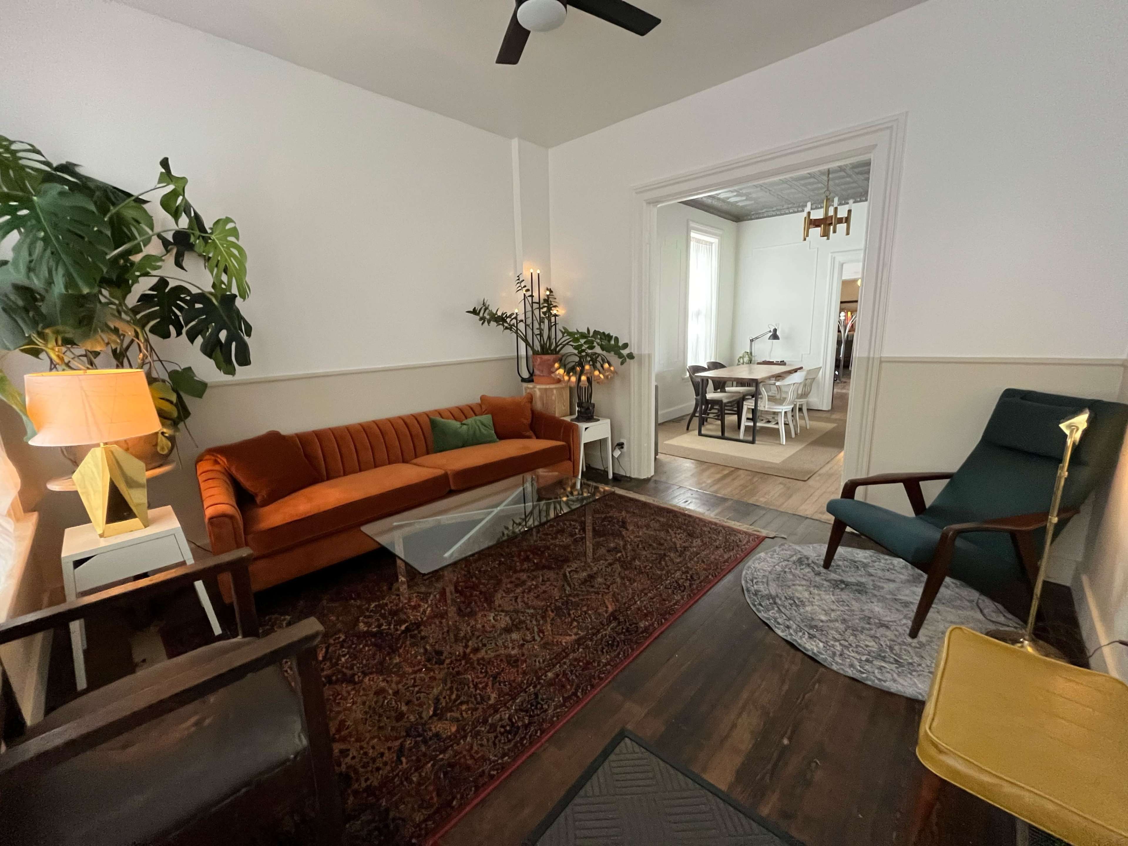 A cozy living room featuring an orange sofa, a glass coffee table, and a green armchair, with plants and decorative lighting adding to the ambiance.