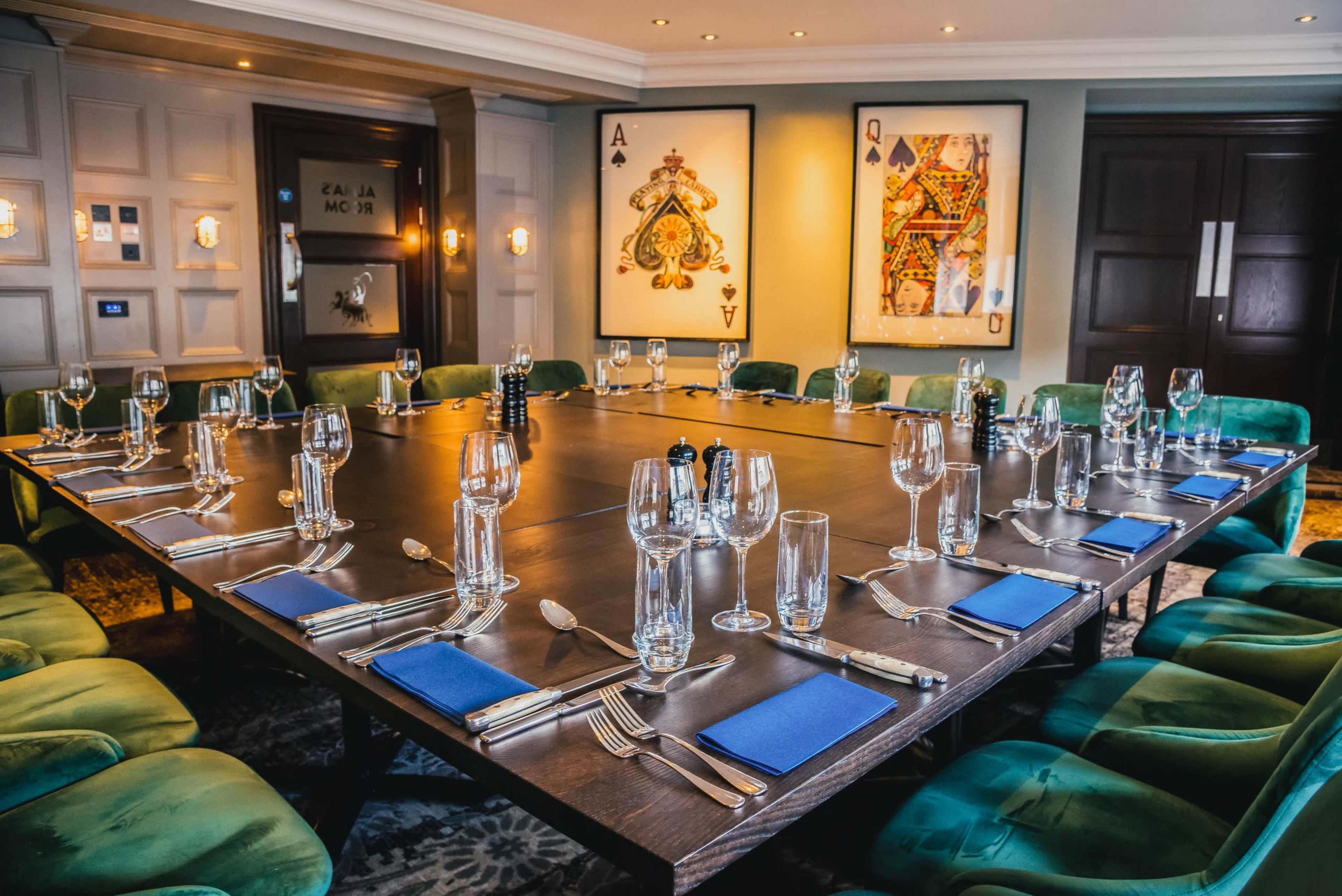 A large dining table is set with glassware, silverware, and blue napkins in a well-decorated room featuring playing card artwork.