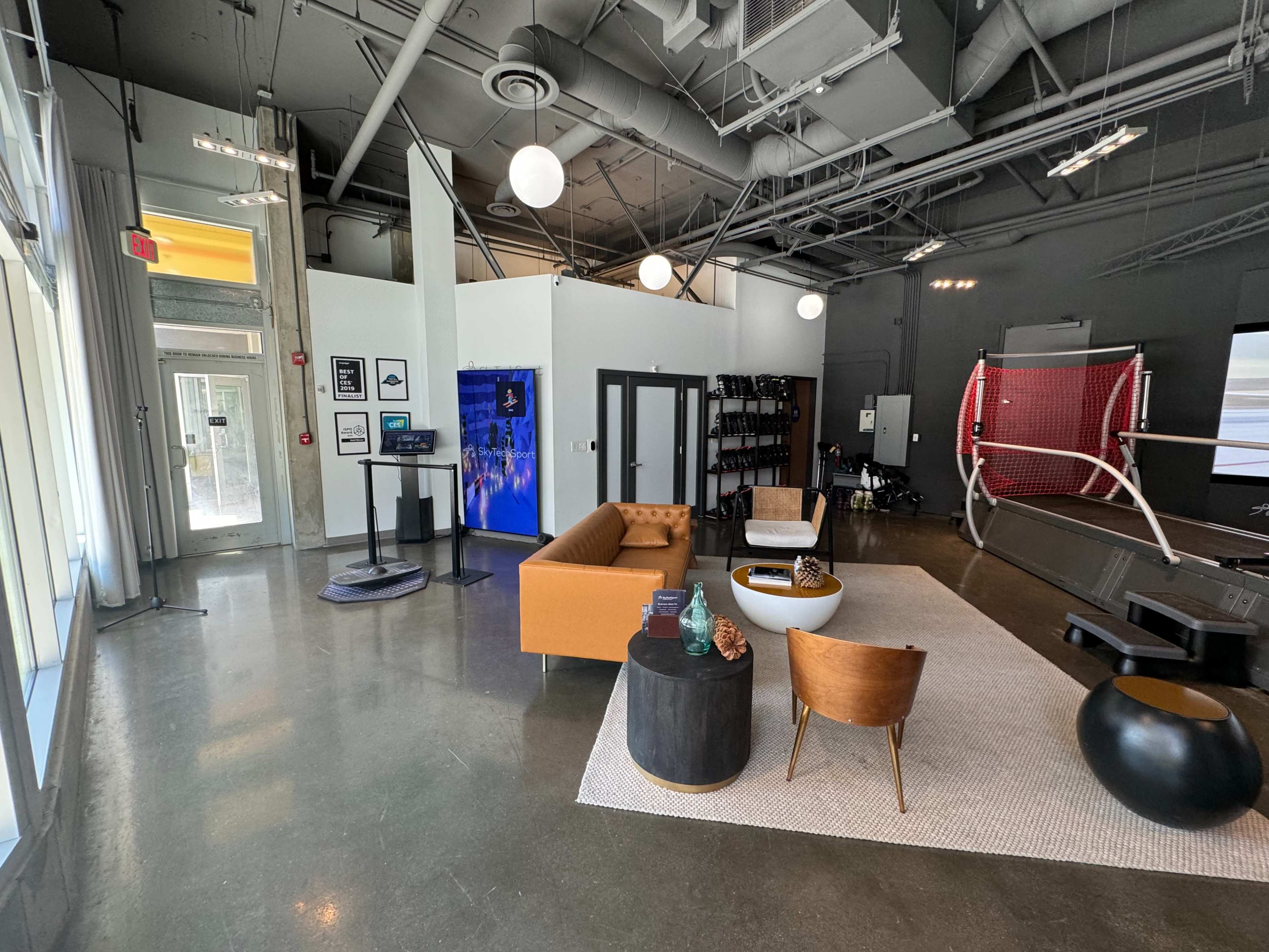 The image shows a modern open space with a seating area featuring a brown sofa and black coffee table, alongside a wall-mounted display and a sports court area in the background.
