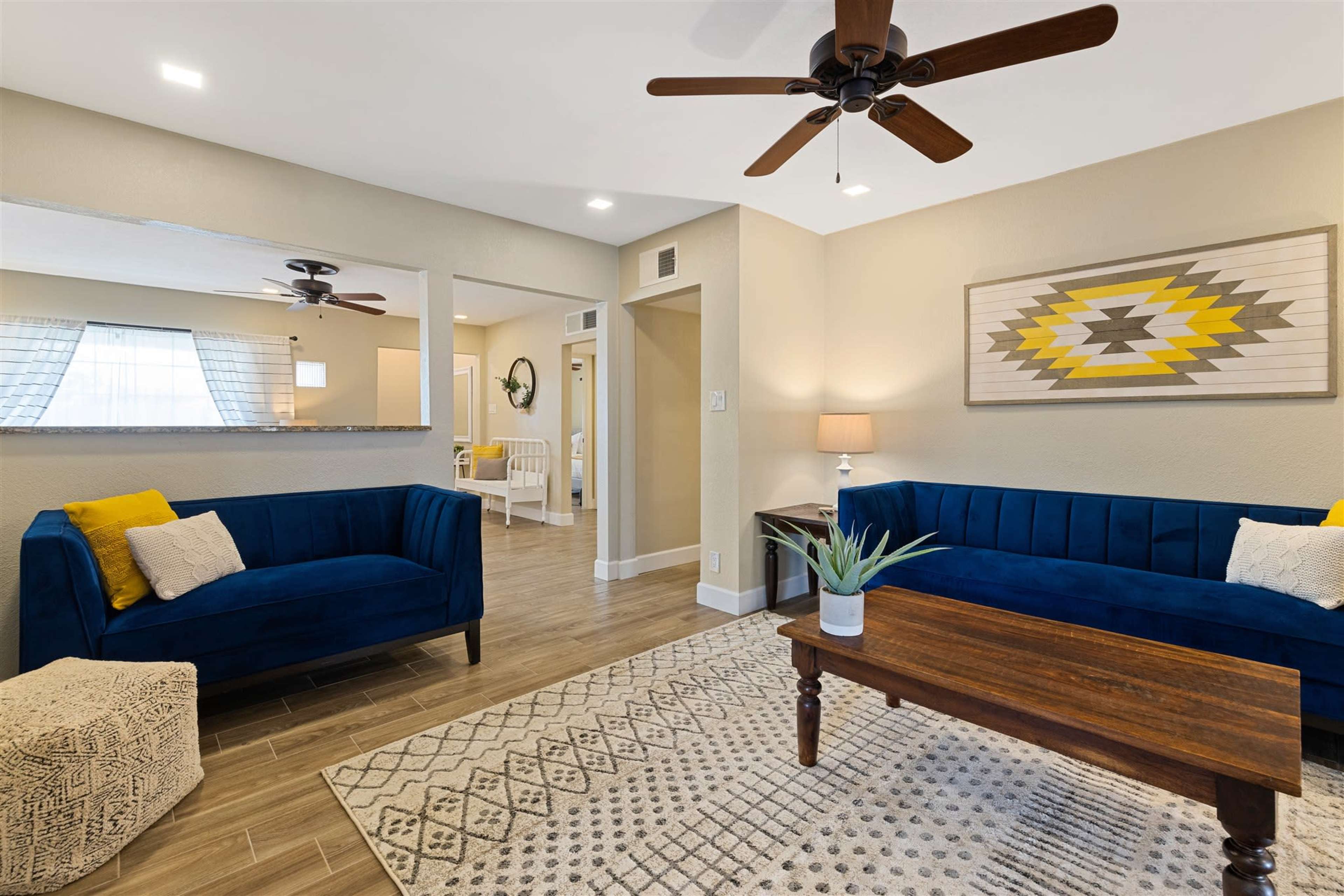 A living room featuring blue sofas, a wooden coffee table, and a patterned rug, with a bright atmosphere enhanced by ceiling fans and ample light.