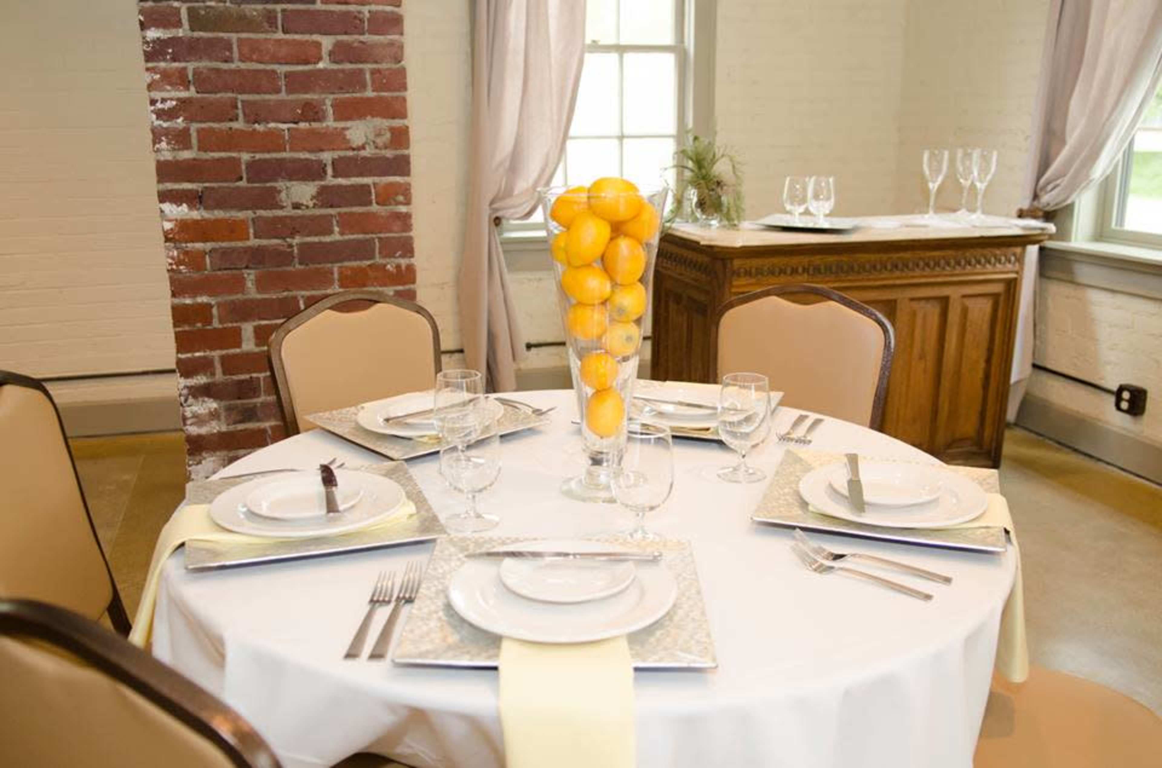 A round dining table is set with white plates, glassware, and a centerpiece of lemons in a clear vase, surrounded by beige chairs in a well-lit room with brick walls.