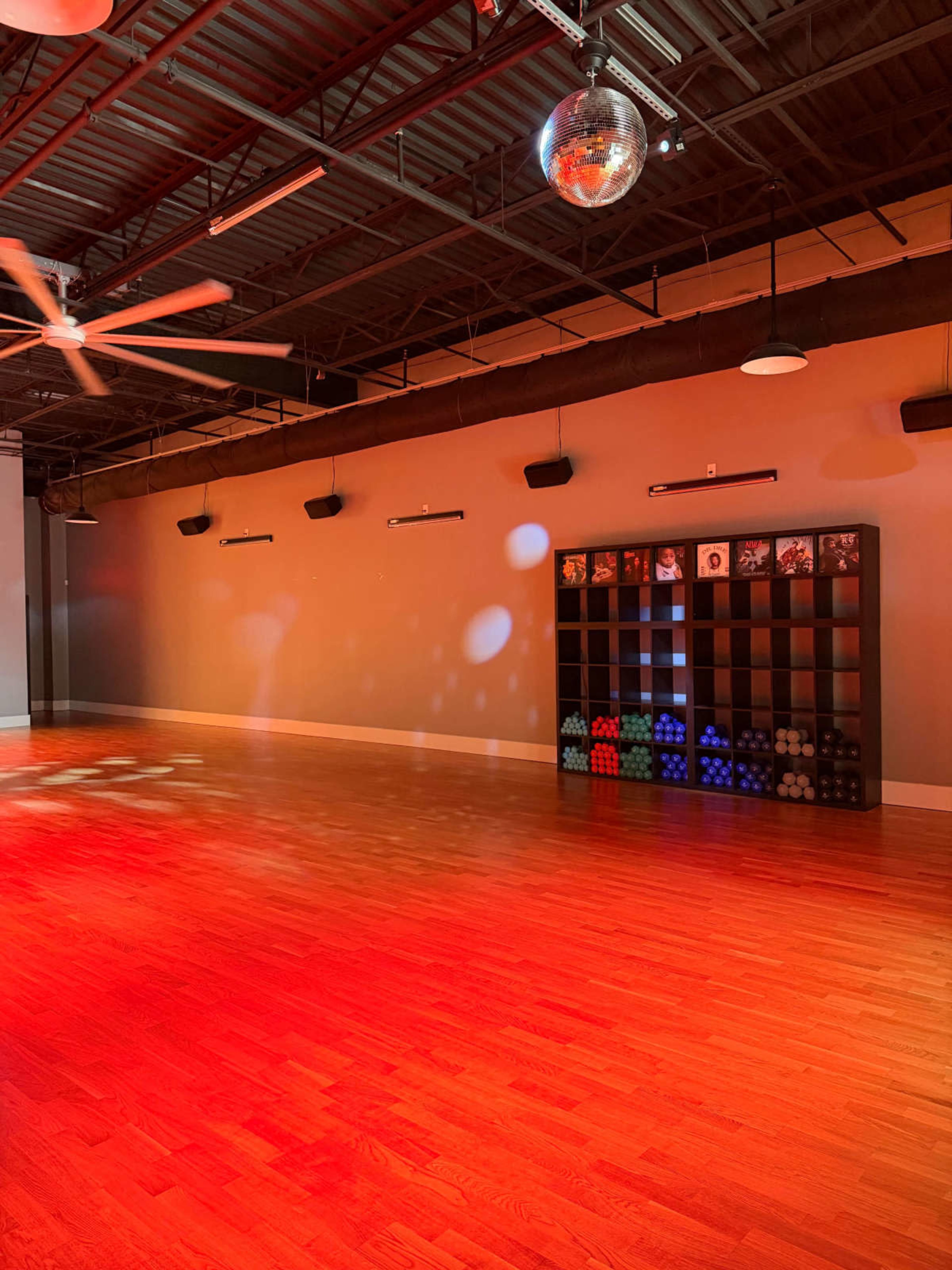 The image shows an empty fitness studio with wooden flooring, a ceiling fan, and a disco ball, illuminated by red lights.
