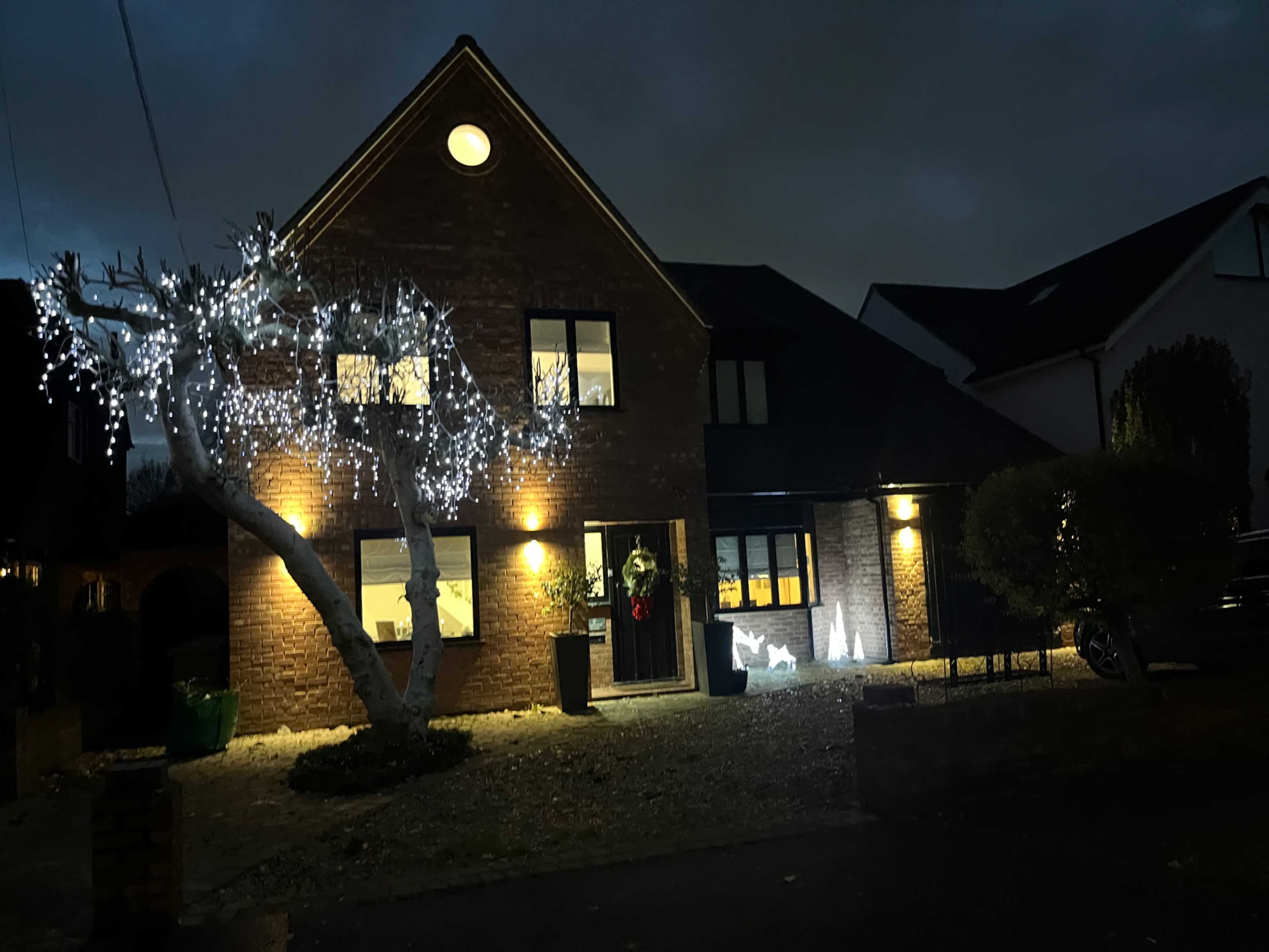 A two-story brick house is illuminated by outdoor lights under a dark sky, with a tree adorned with white lights in the front yard.
