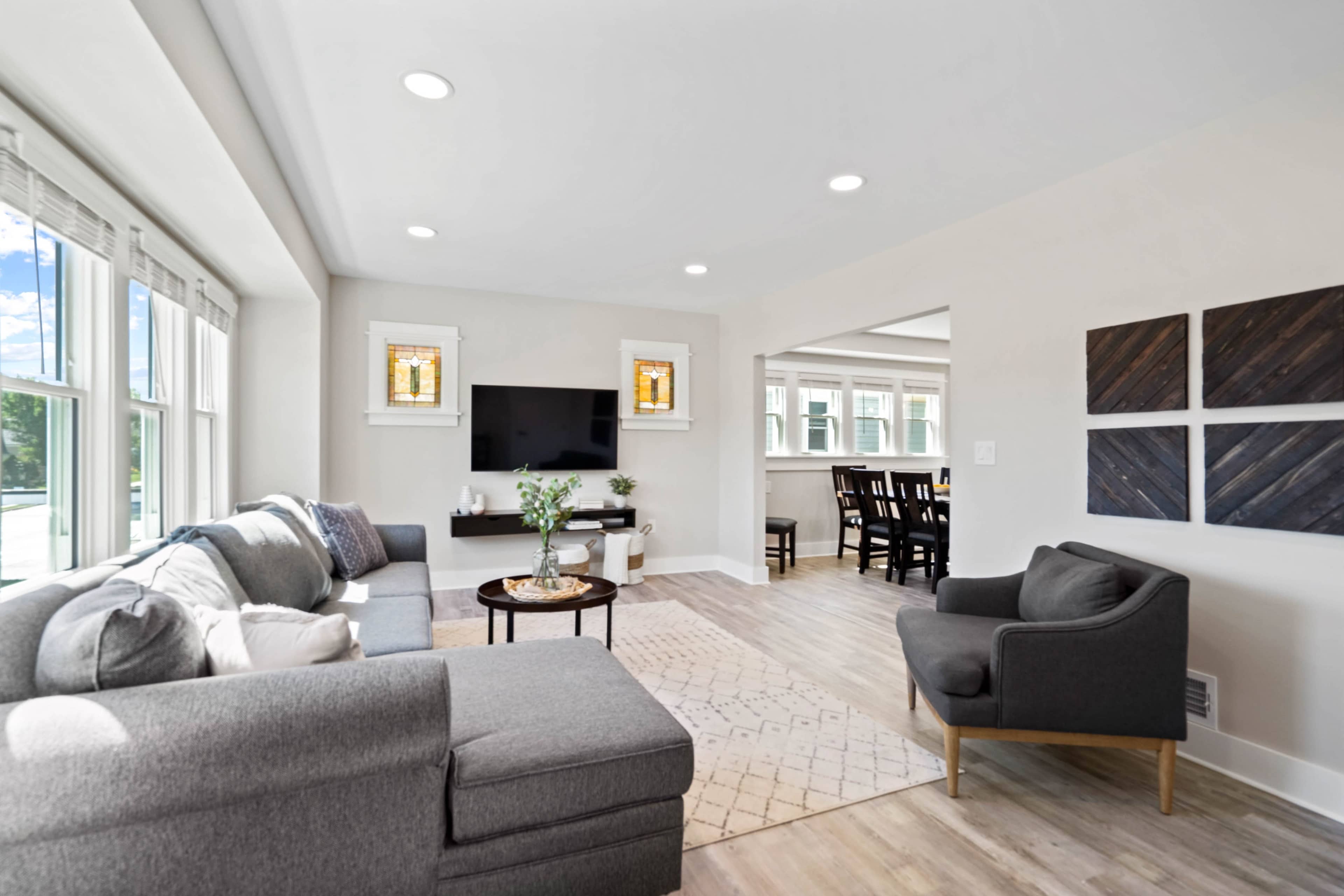 A modern living room features a gray sectional sofa, a round coffee table, and a TV mounted on the wall, with clear windows allowing natural light.