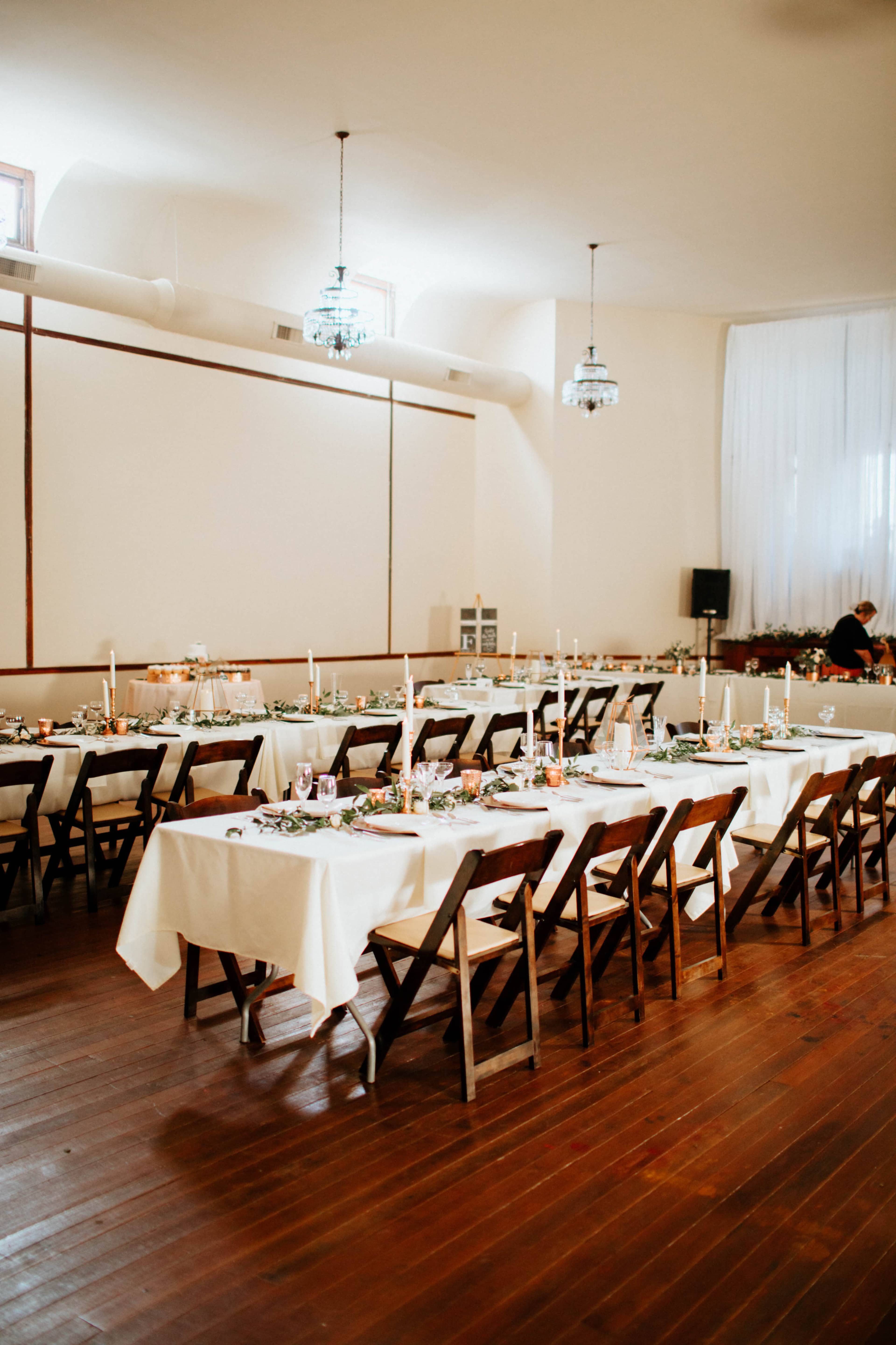 A spacious room features rows of long tables set with white tablecloths and candles, surrounded by wooden chairs.