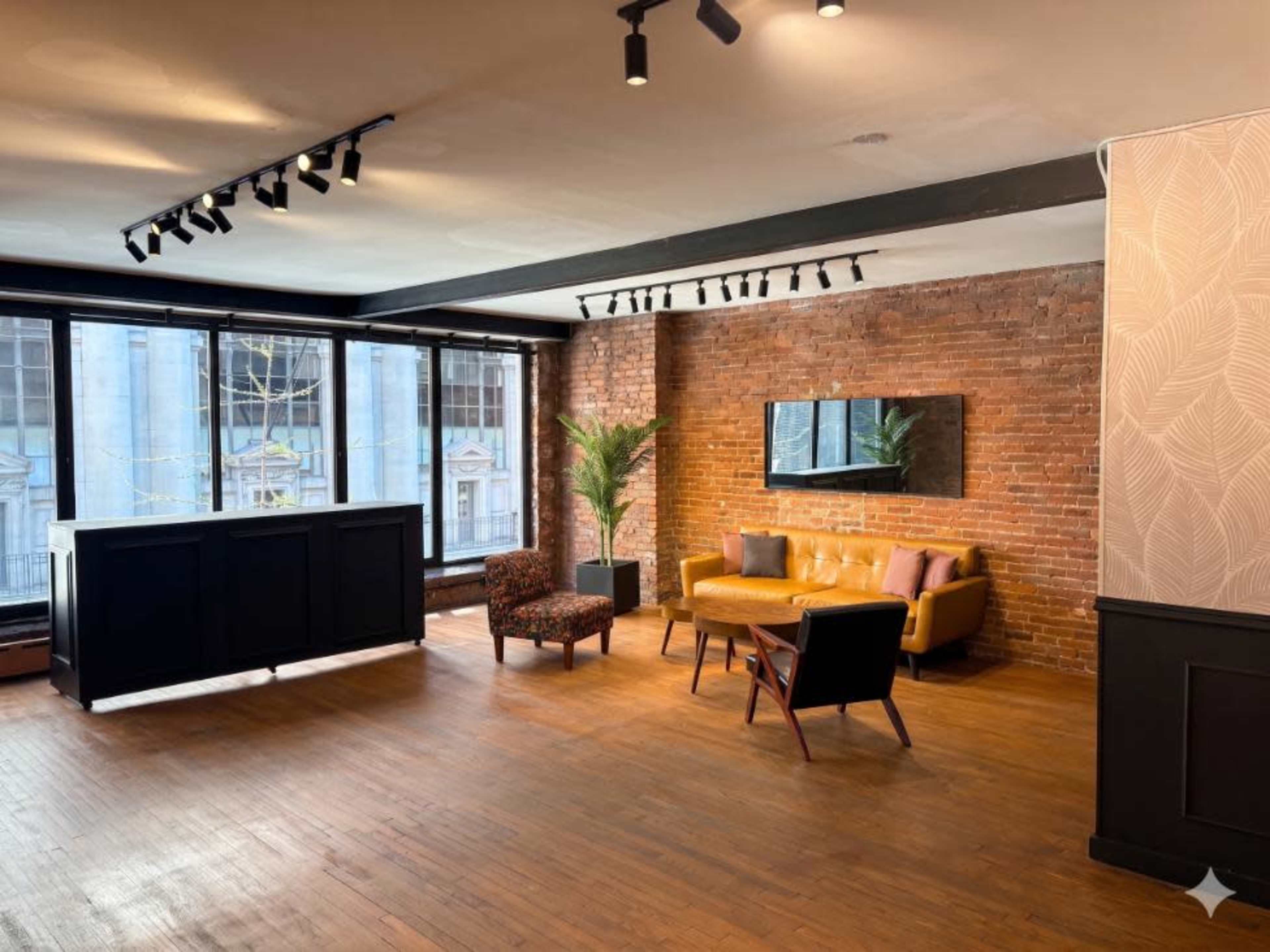 The image shows a spacious interior with exposed brick walls, large windows, and a seating area featuring a yellow sofa, a round wooden coffee table, and a patterned armchair.