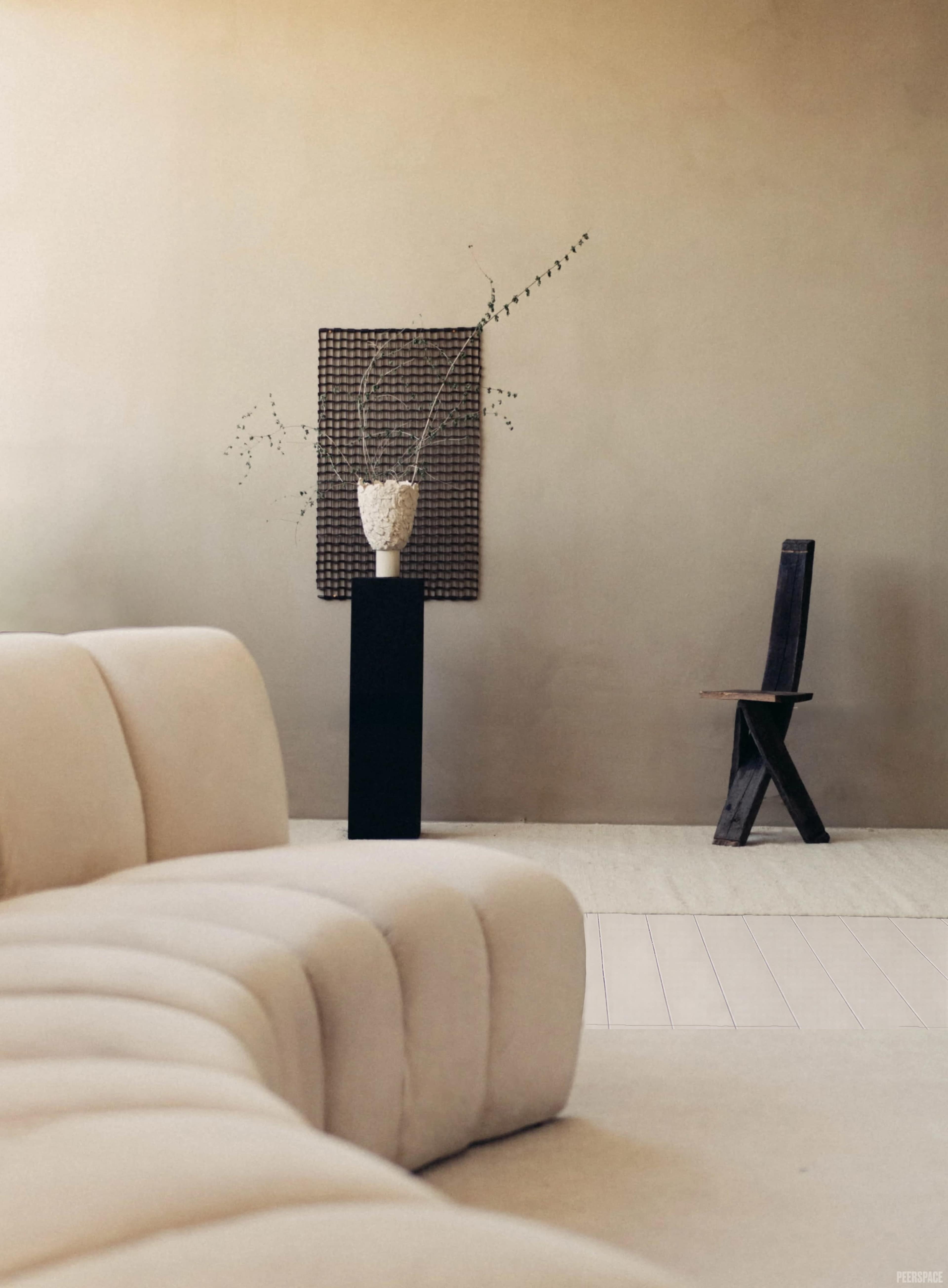 A minimalist interior features a curved sofa, a tall black pedestal with a vase of branches, and a wooden chair against a neutral wall.