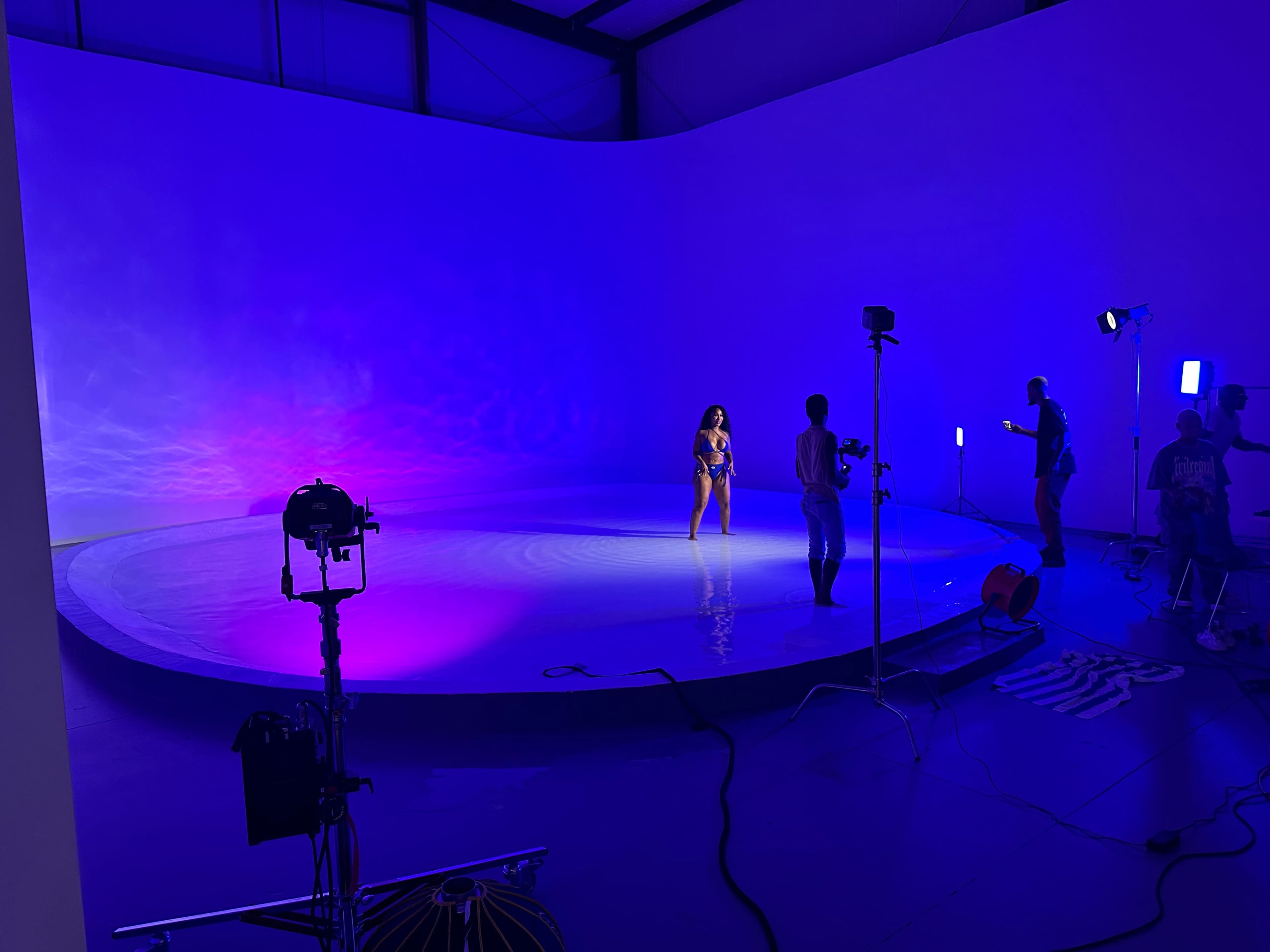 A model poses on a circular platform surrounded by colorful lights while a crew sets up for a photoshoot in a large studio space.