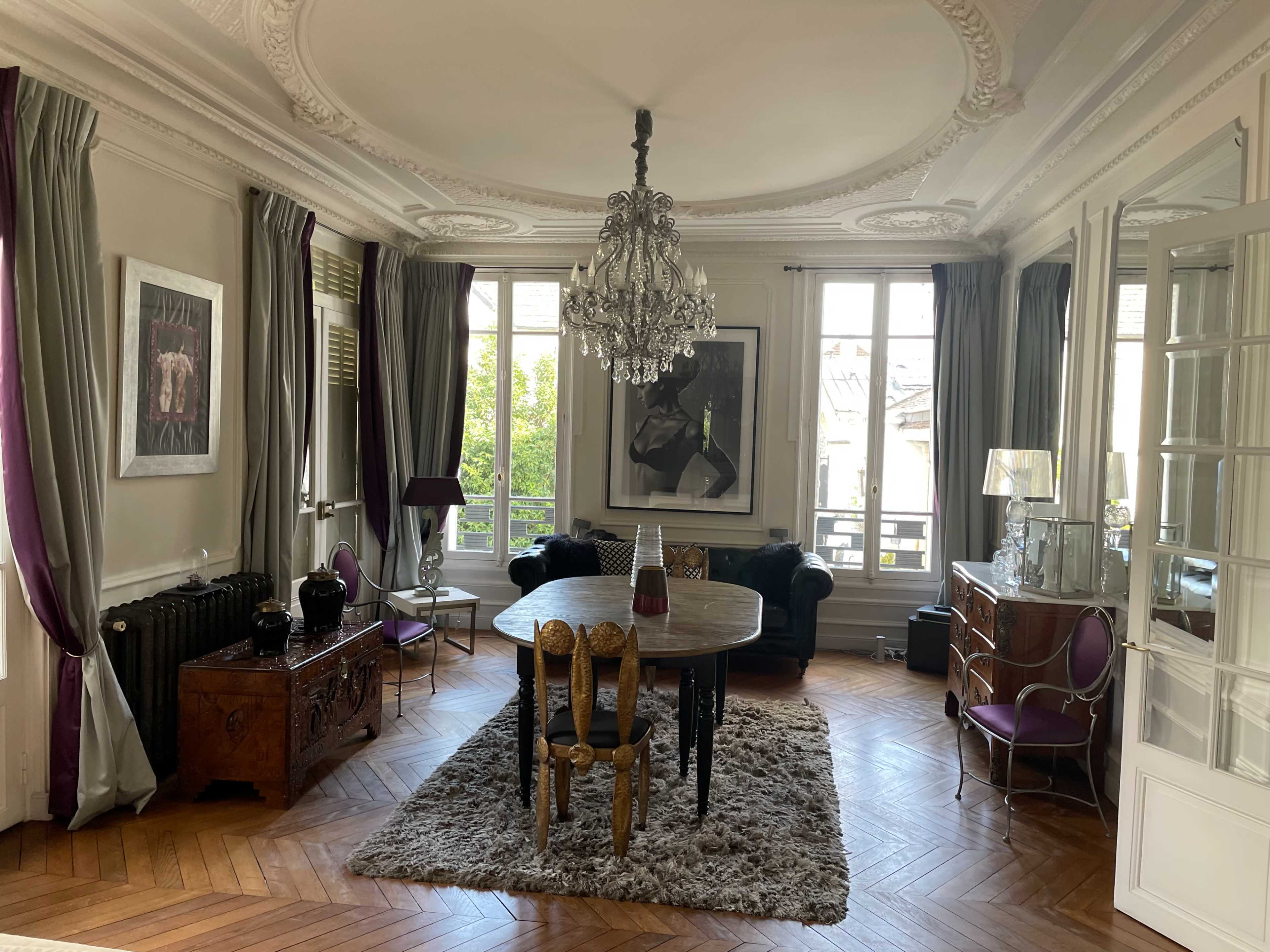 A spacious living room features a round table, a chandelier, and large windows that overlook an outdoor view.
