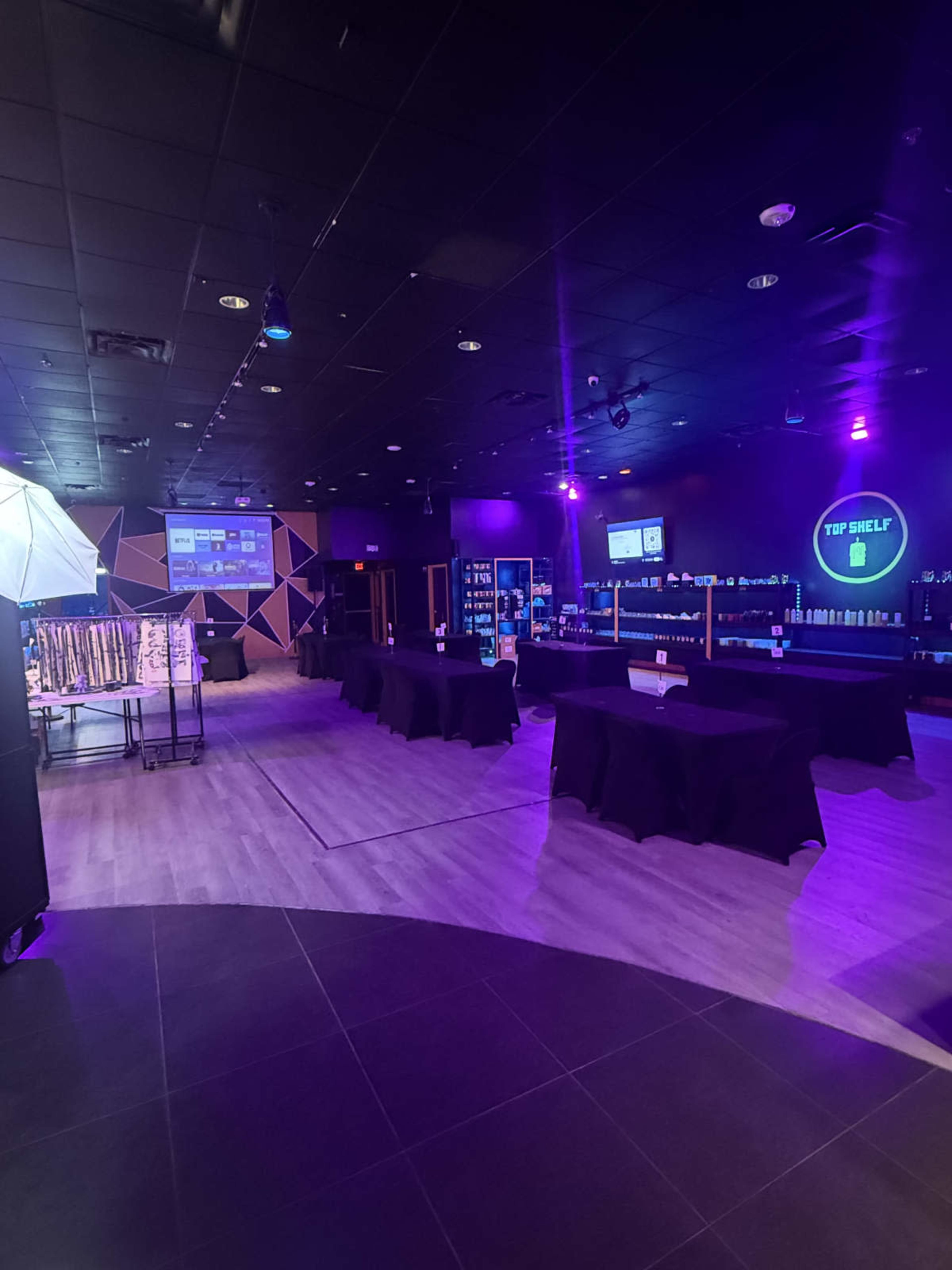 The image depicts a spacious interior of a modern retail space with tables covered in black cloth, illuminated by purple lighting and featuring various displays against the walls.