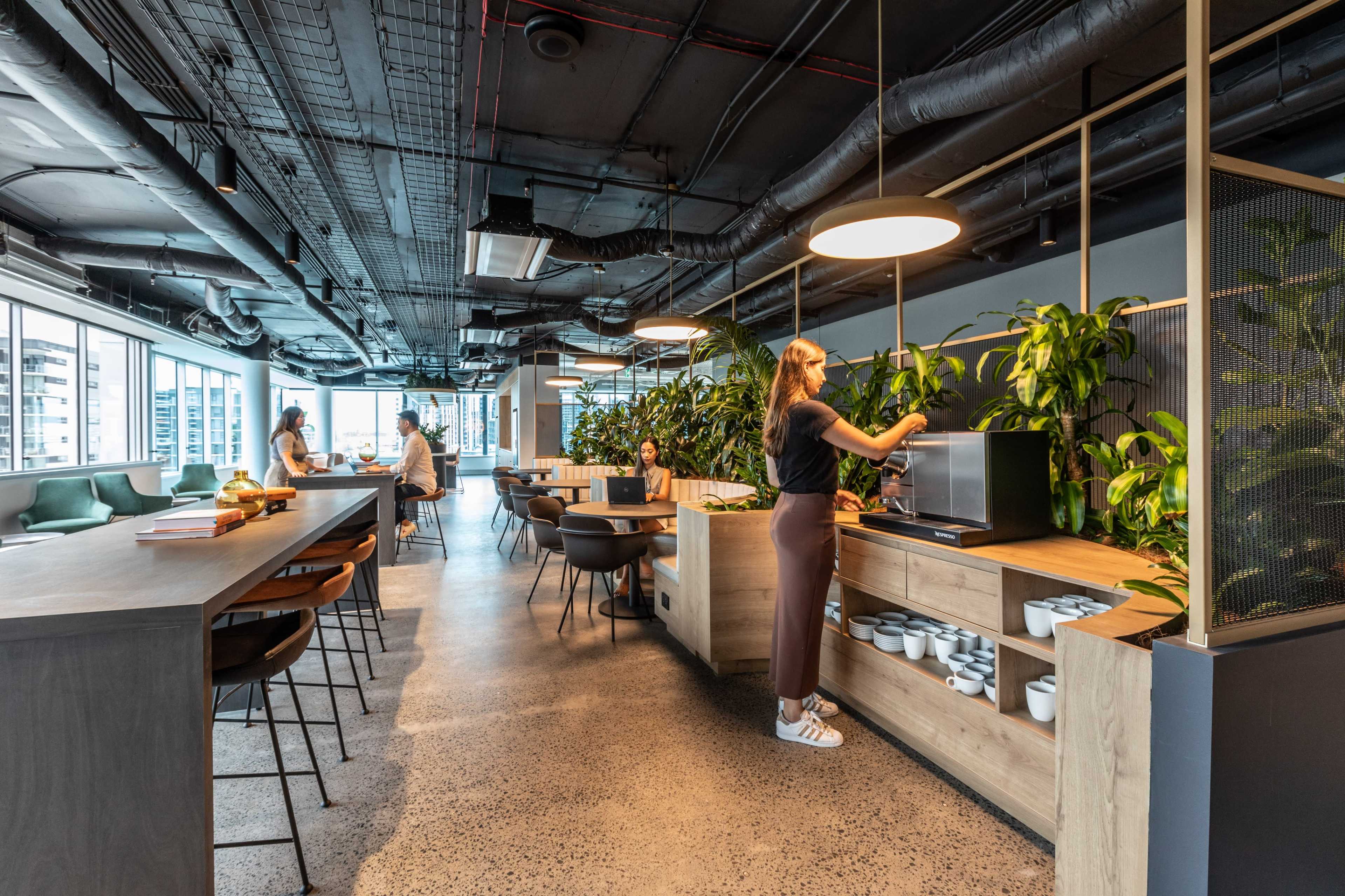 A woman adjusts a microwave in a modern office kitchen area filled with plants and communal seating.