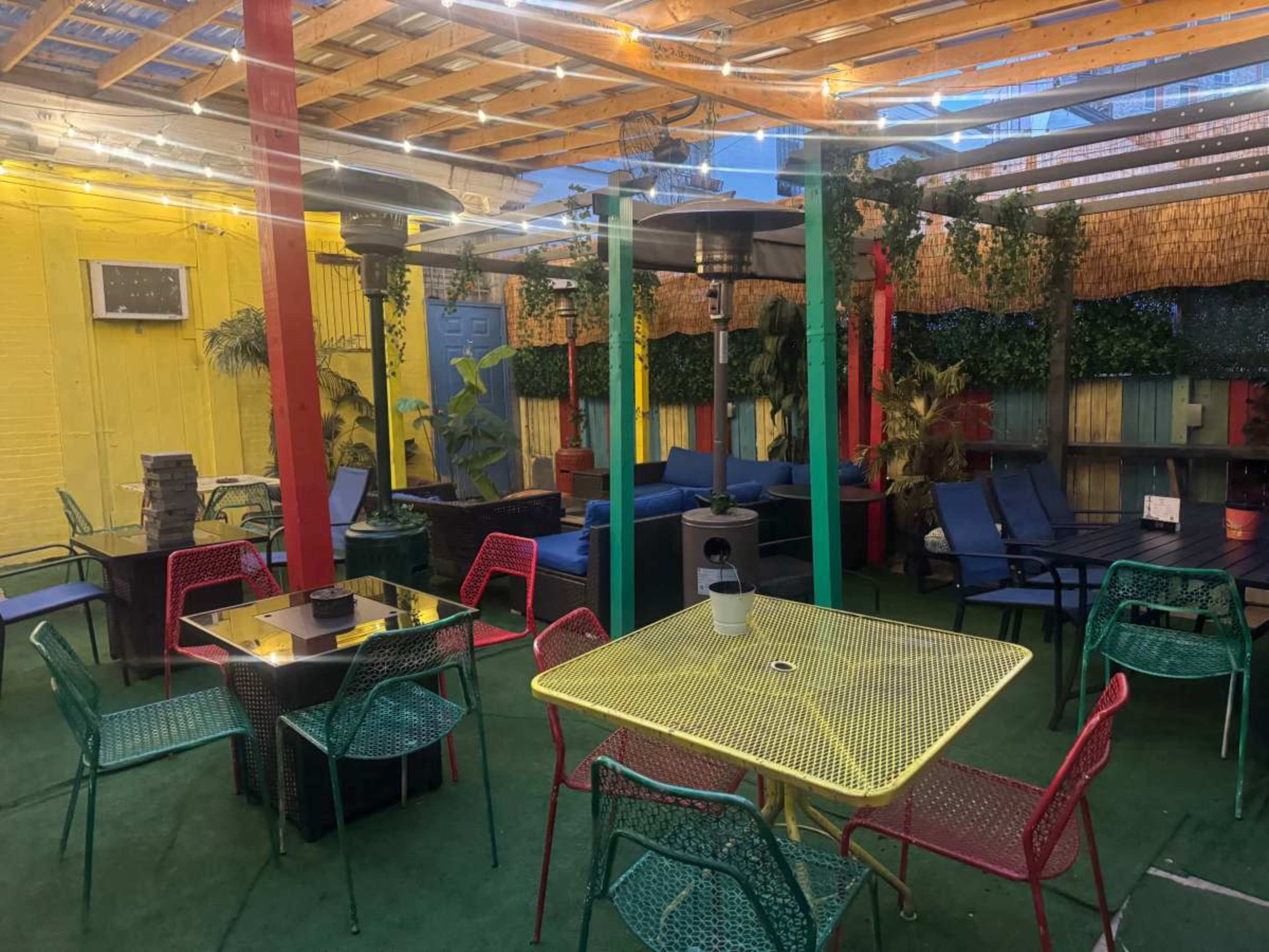 The image shows a colorful outdoor seating area with multicolored chairs and tables, surrounded by plants and decorative lighting.