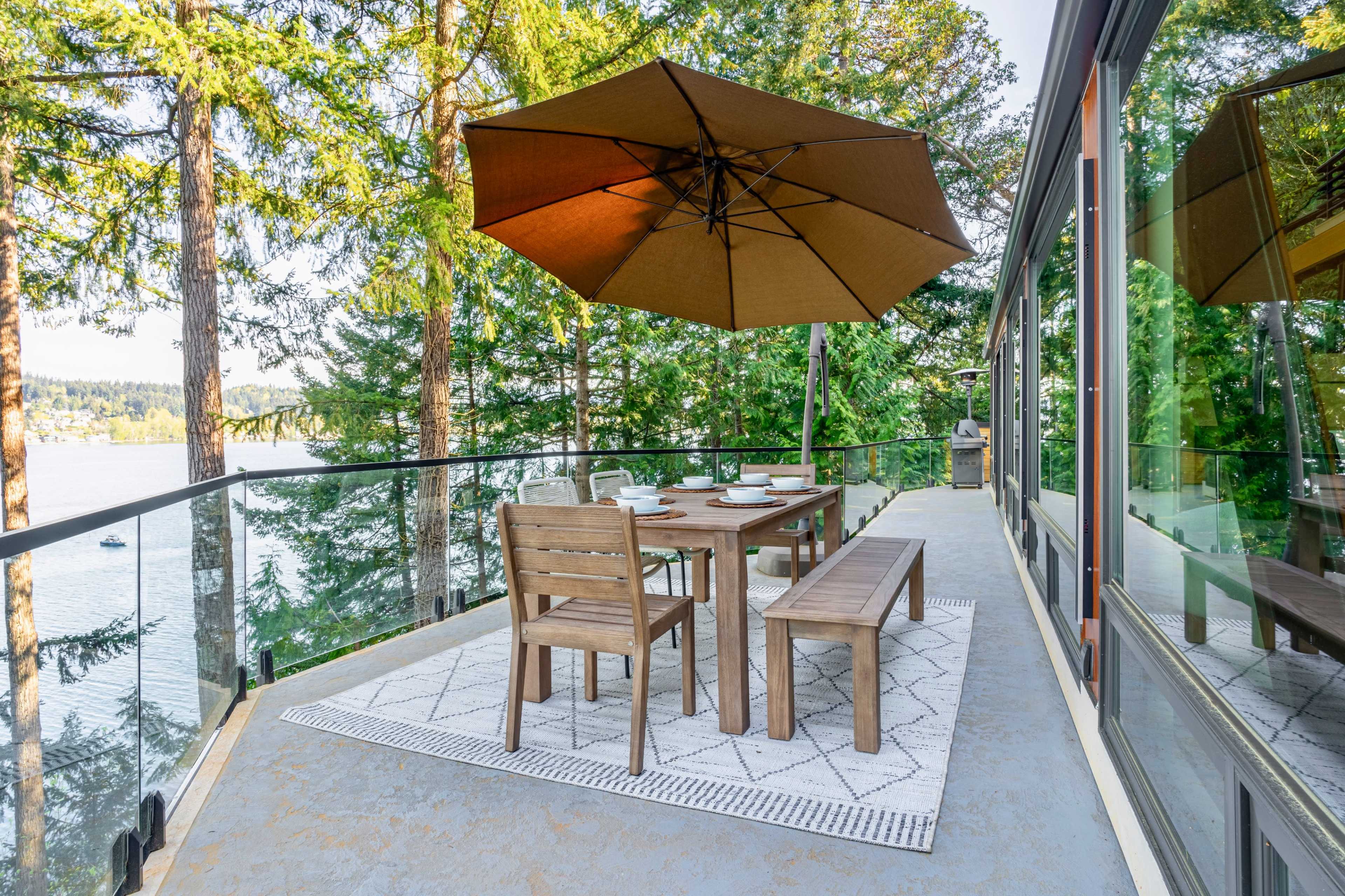 A wooden dining set with an umbrella is positioned on a balcony overlooking a body of water, surrounded by tall trees.