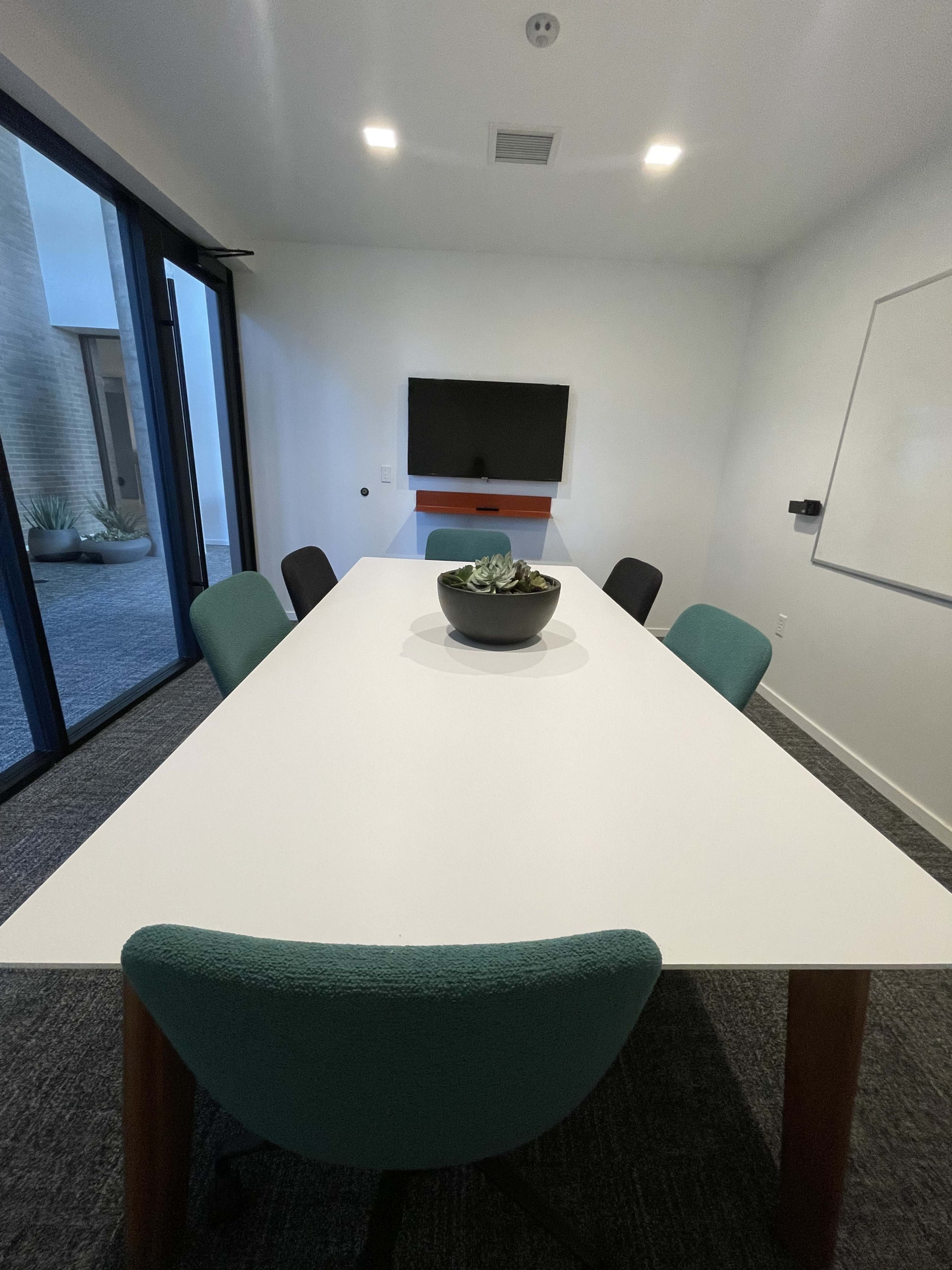 A conference room features a long white table surrounded by six chairs and includes a wall-mounted television and a whiteboard.
