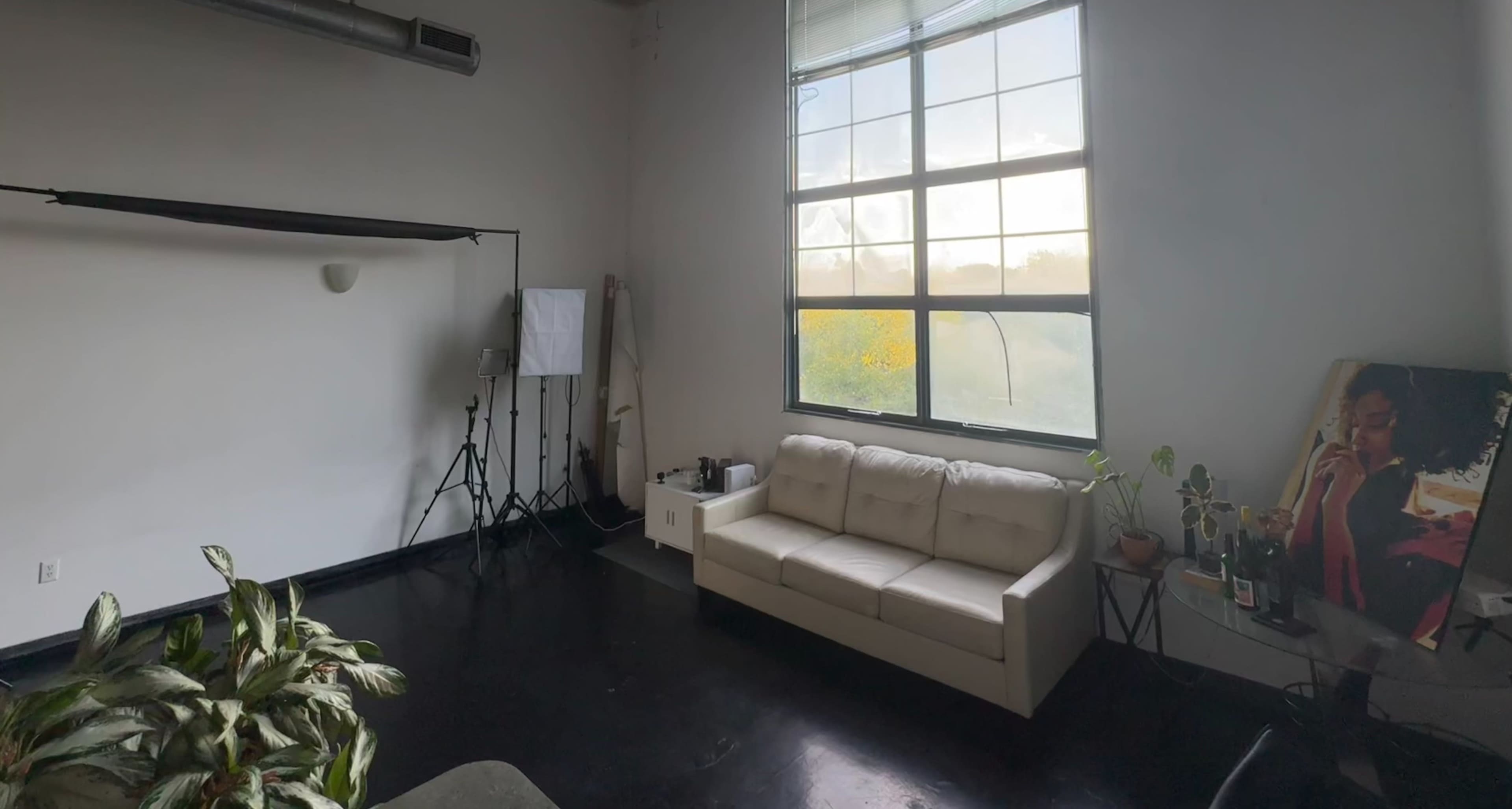 The image shows a spacious room with a large window, a white sofa, and photography equipment in the corner.