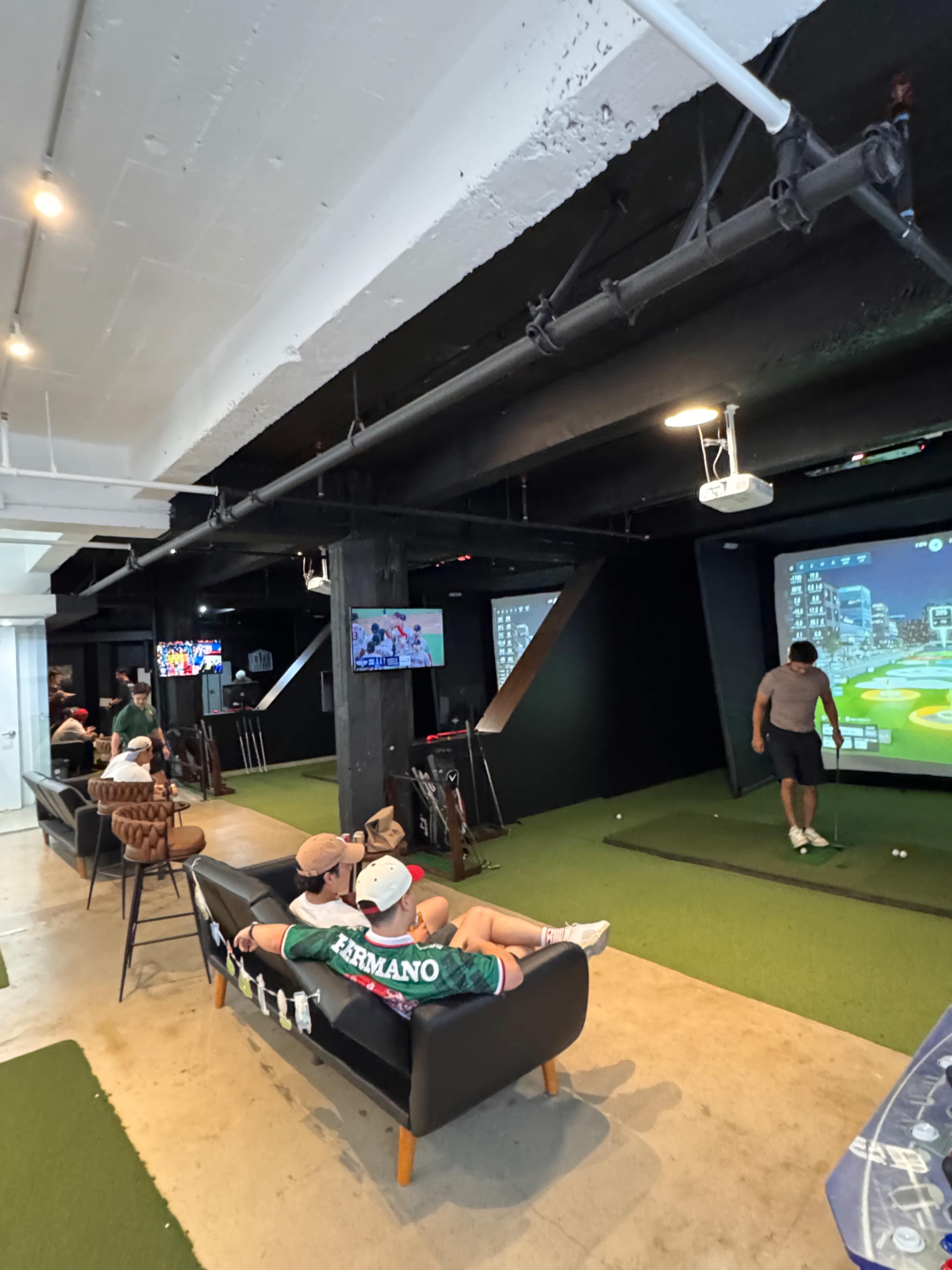 The image shows a modern indoor space with a golf simulator, two television screens, and several people sitting on a couch.