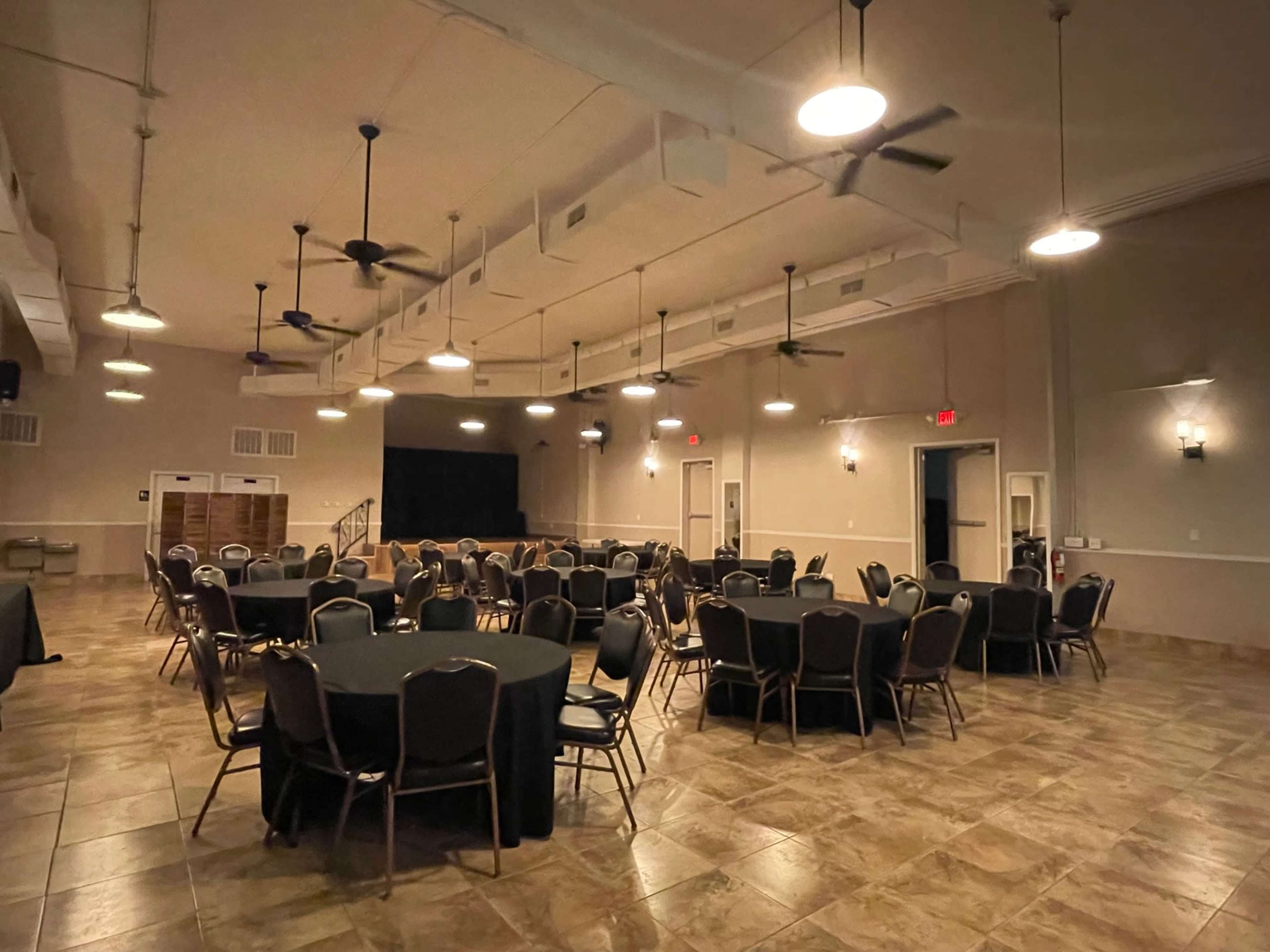 A spacious event room features neatly arranged round tables with black tablecloths and ceiling fans above, illuminated by multiple ceiling lights.