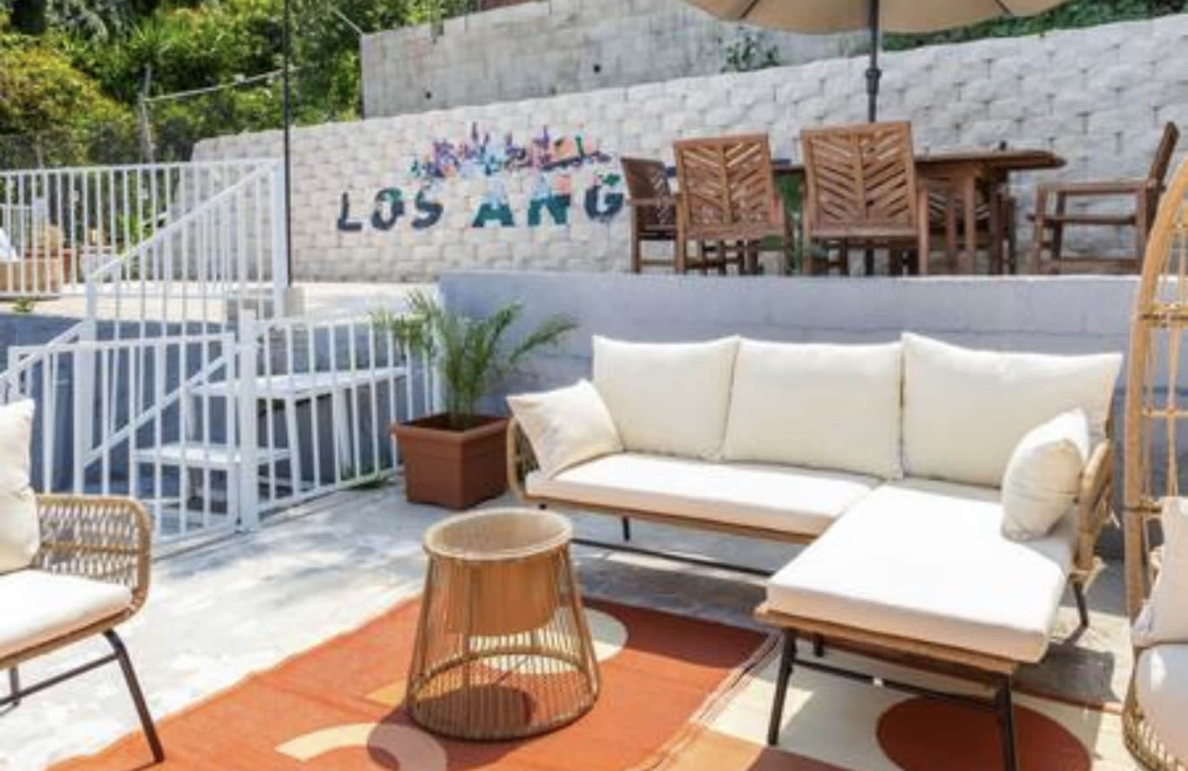 The image shows a patio area with a cream-colored sofa set, a round coffee table, and a wooden dining table in the background, all set against a wall decorated with the words "LOS ANG."