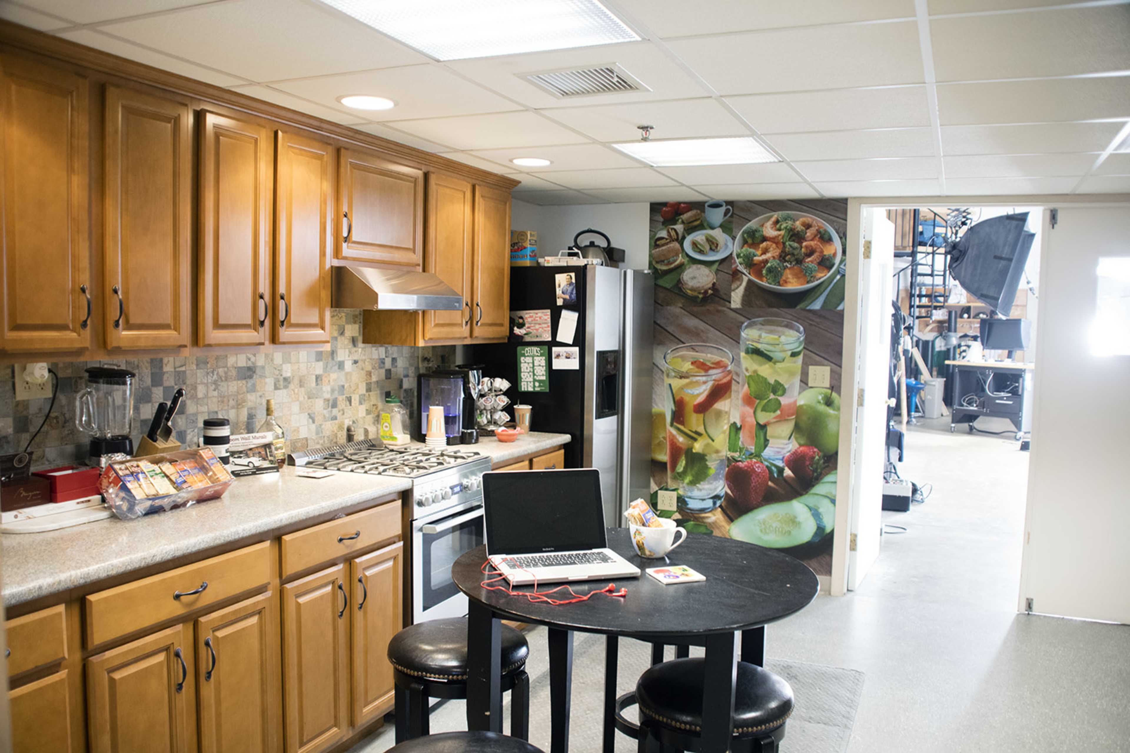 A modern kitchen features wooden cabinets, a stove, and a table with a laptop, adjacent to a room with a colorful food-themed mural and studio equipment.