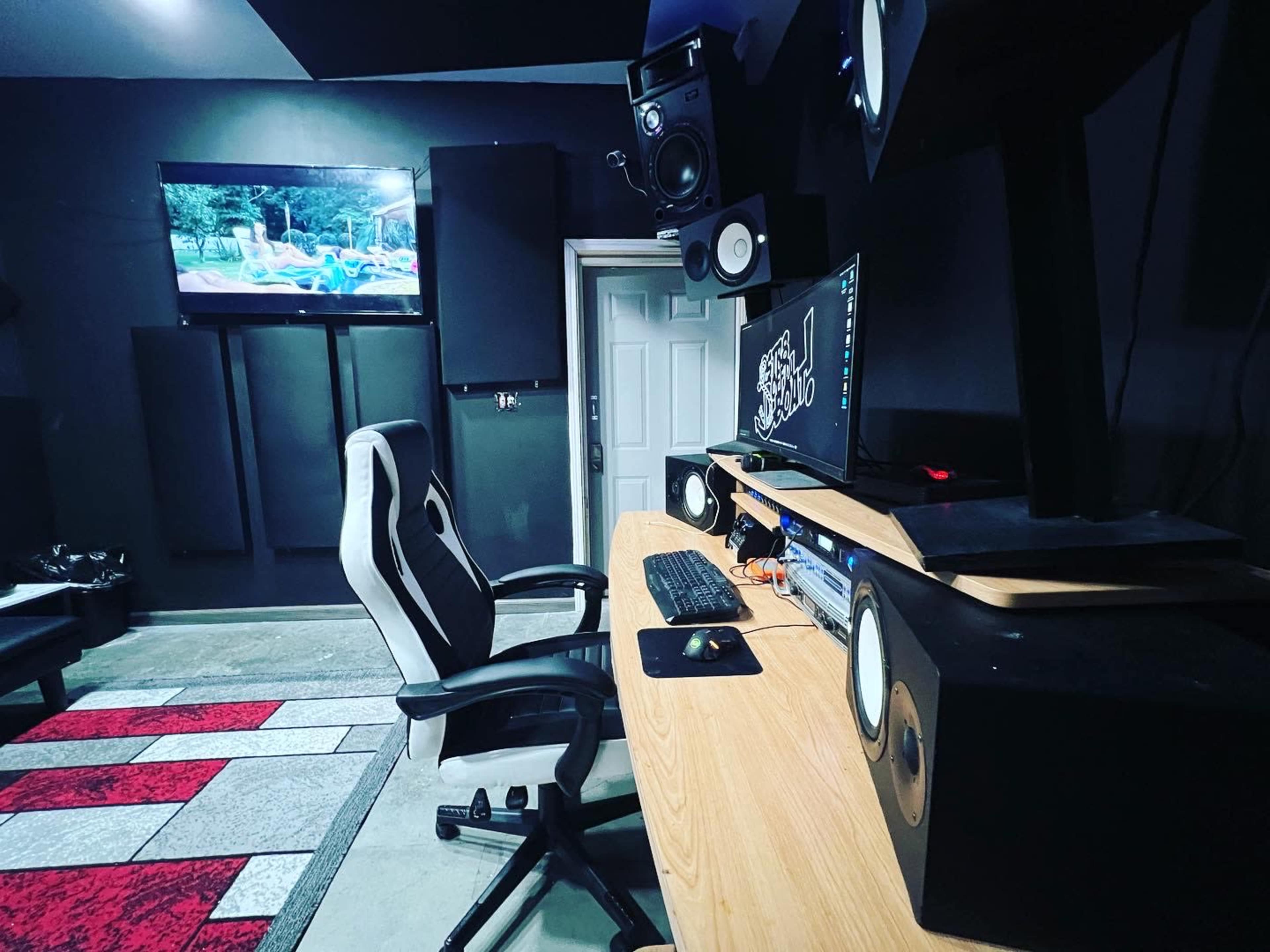 The image shows a recording studio with a wooden desk, a computer setup, large speakers, and a television mounted on the wall.