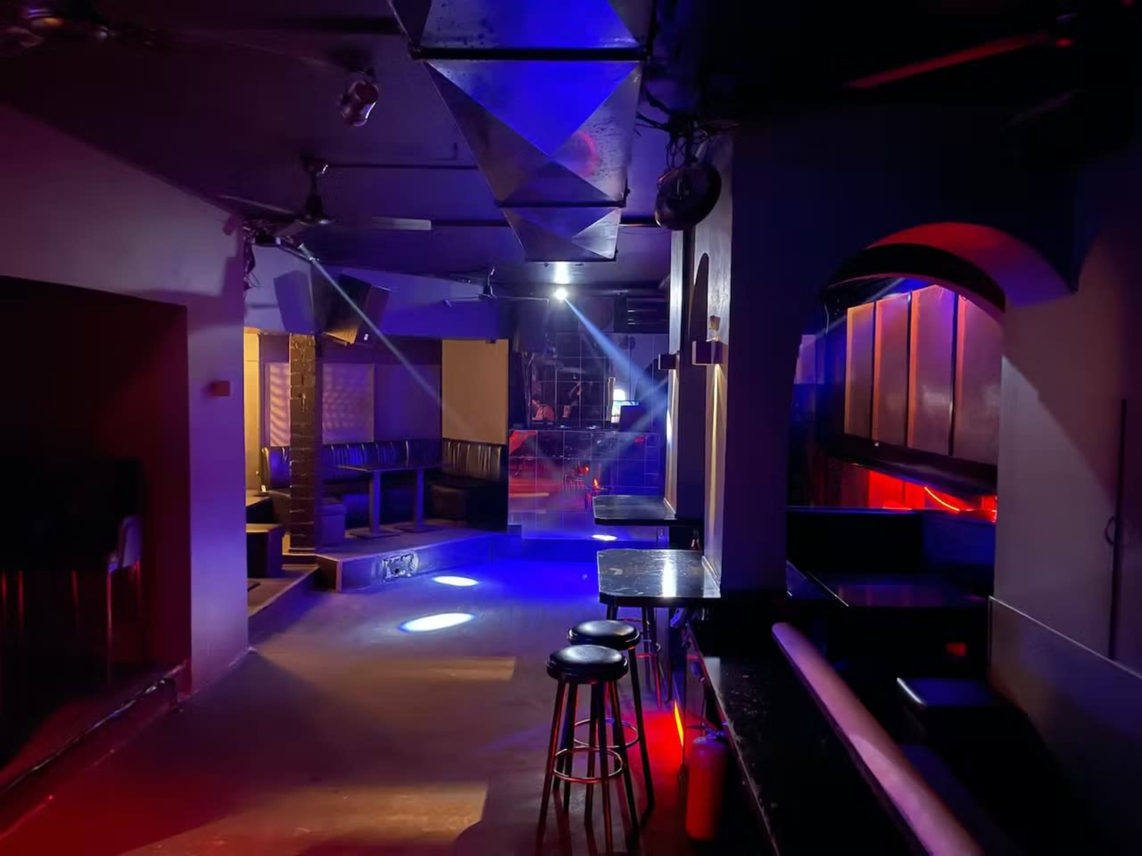 A dimly lit nightclub interior features empty seating areas, polished surfaces, and colorful lighting effects.