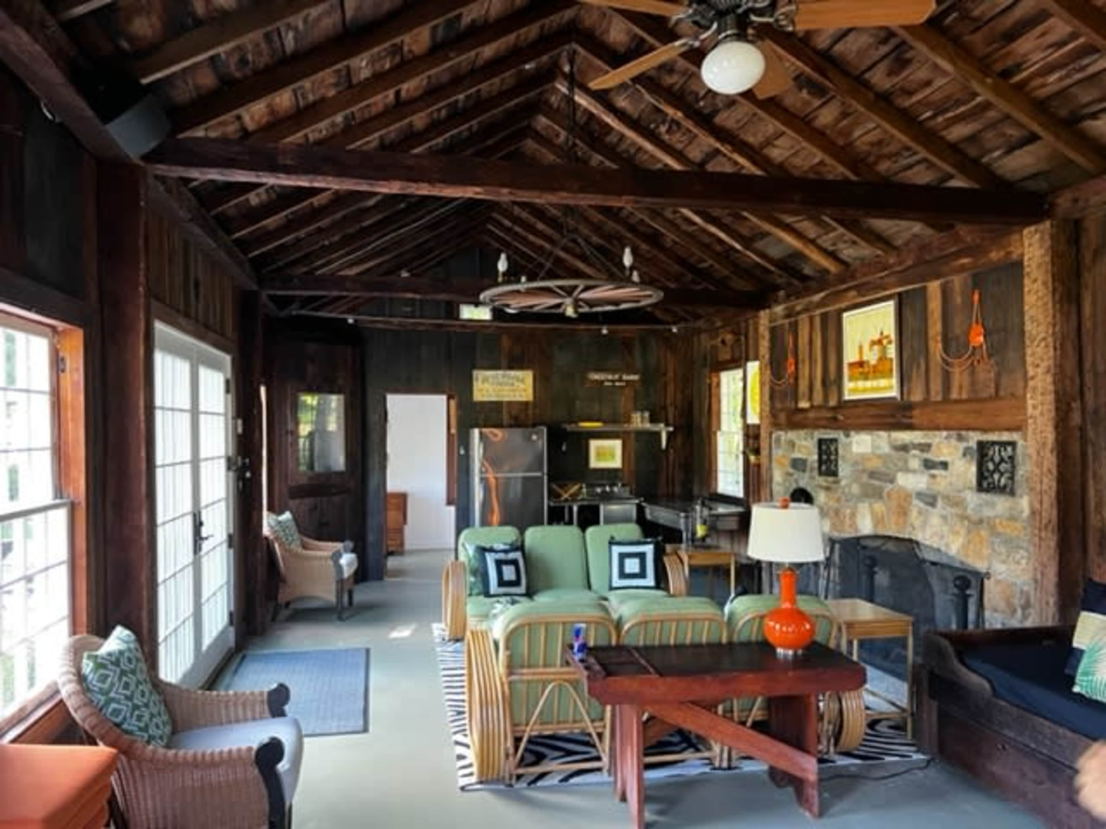 The interior of a rustic cabin features exposed wooden beams, a stone fireplace, and a mix of seating arrangements including chairs and a sofa.