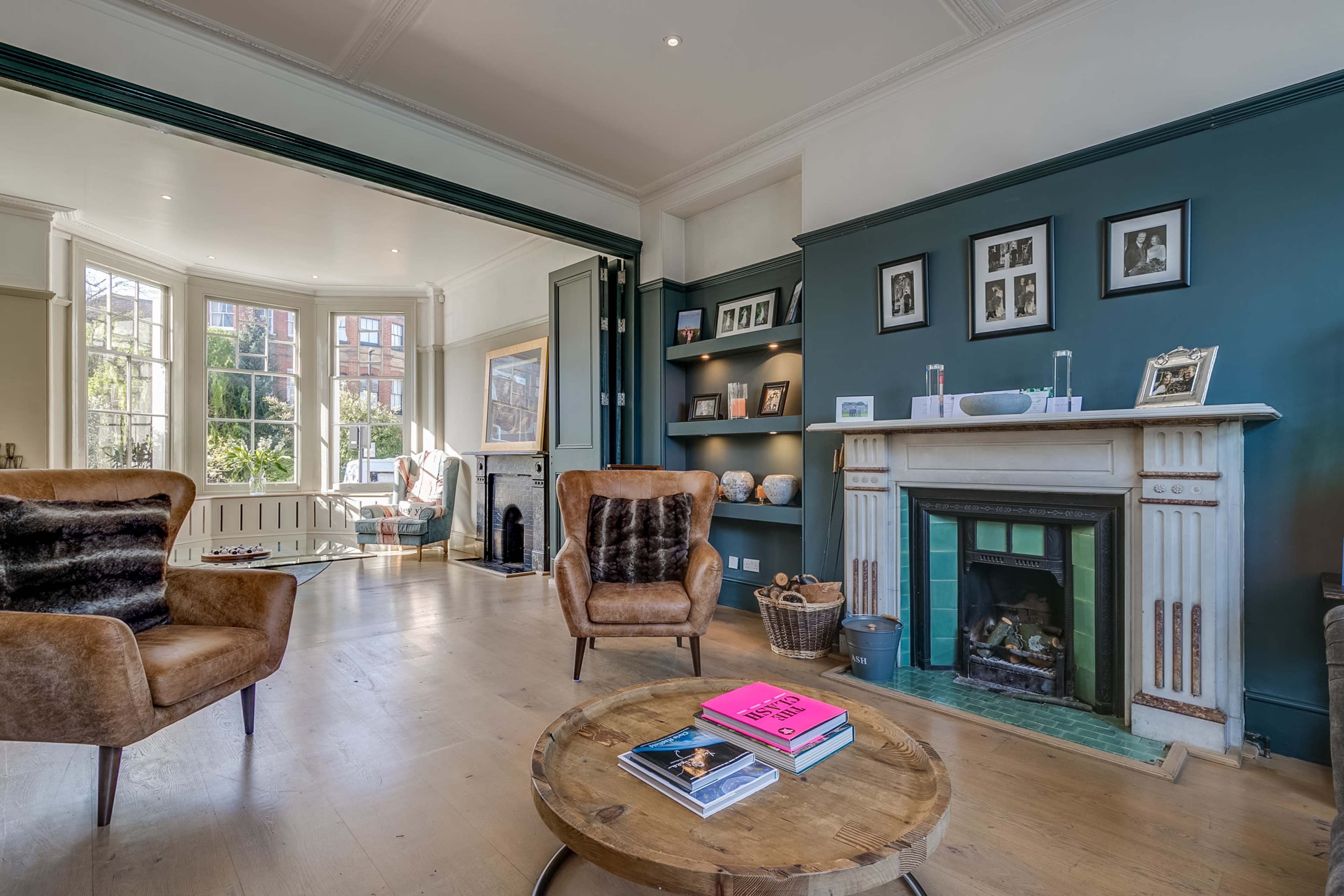 A living room with two brown armchairs facing a circular wooden coffee table, a fireplace with a green surround, and bookshelves featuring photographs and decorative items.