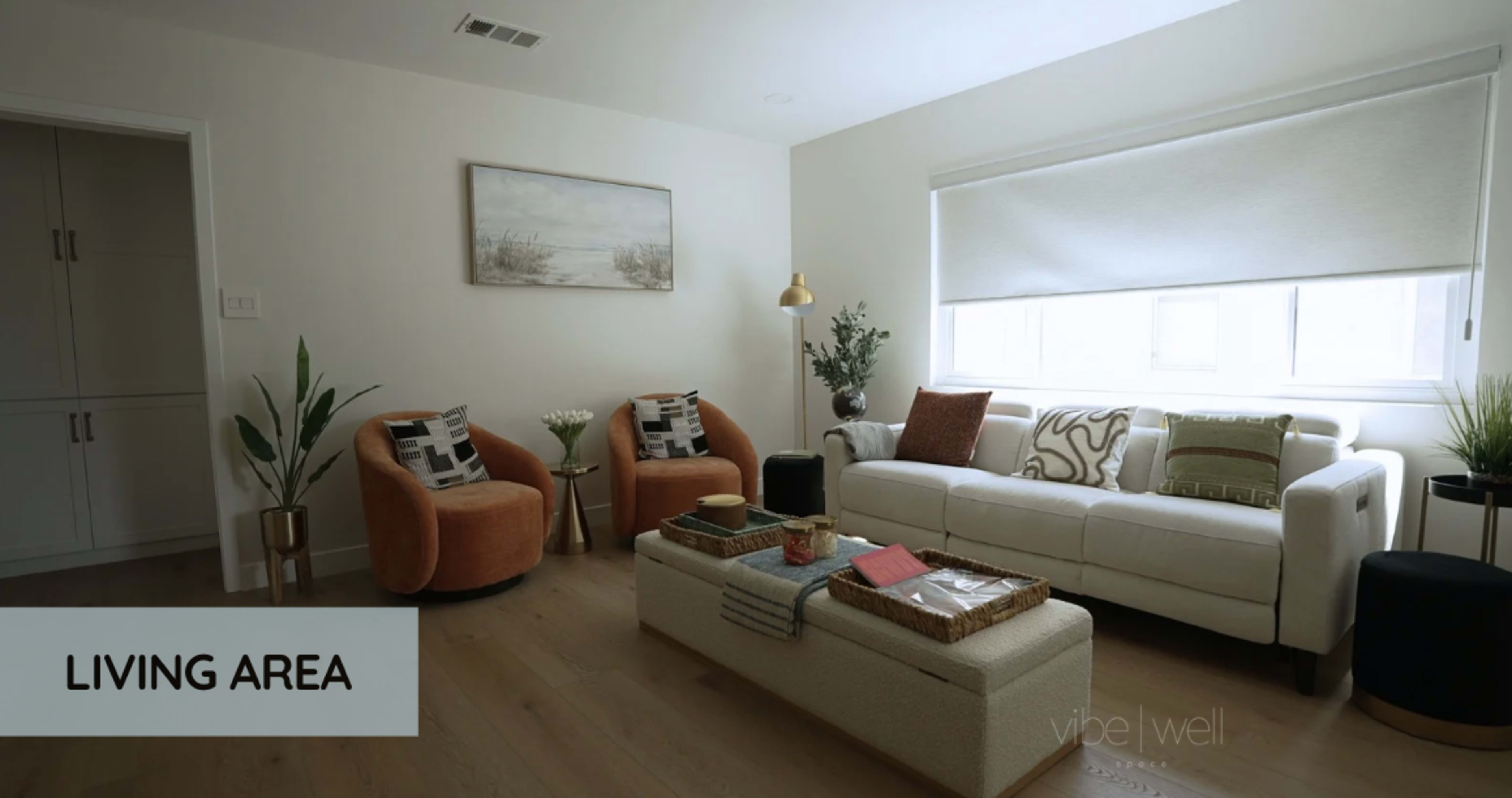 The image shows a modern living area with a white couch, two orange armchairs, a coffee table, and large windows covered by light shades.