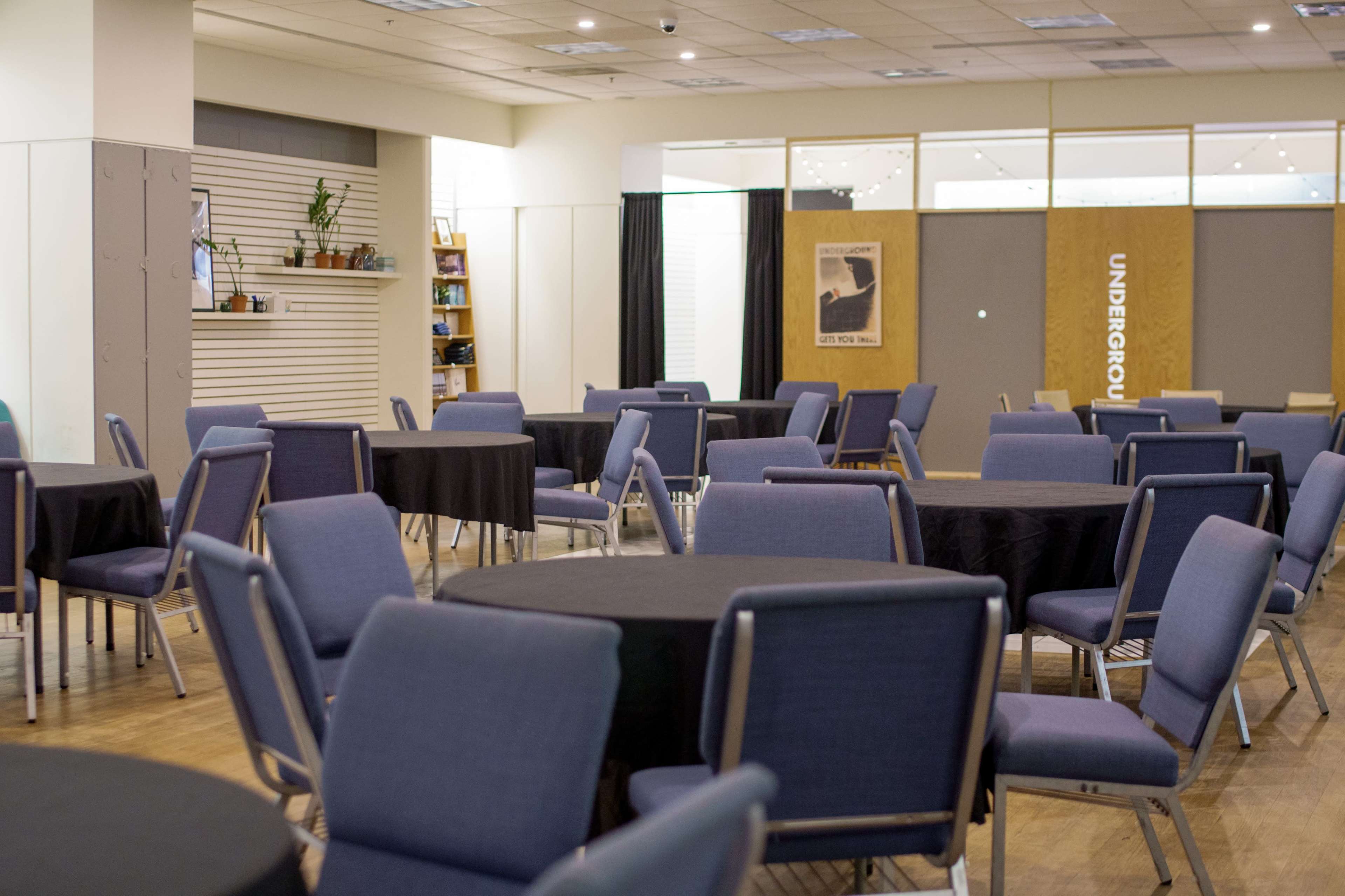 The image shows a spacious indoor room with several round tables covered in black tablecloths and surrounded by blue chairs.