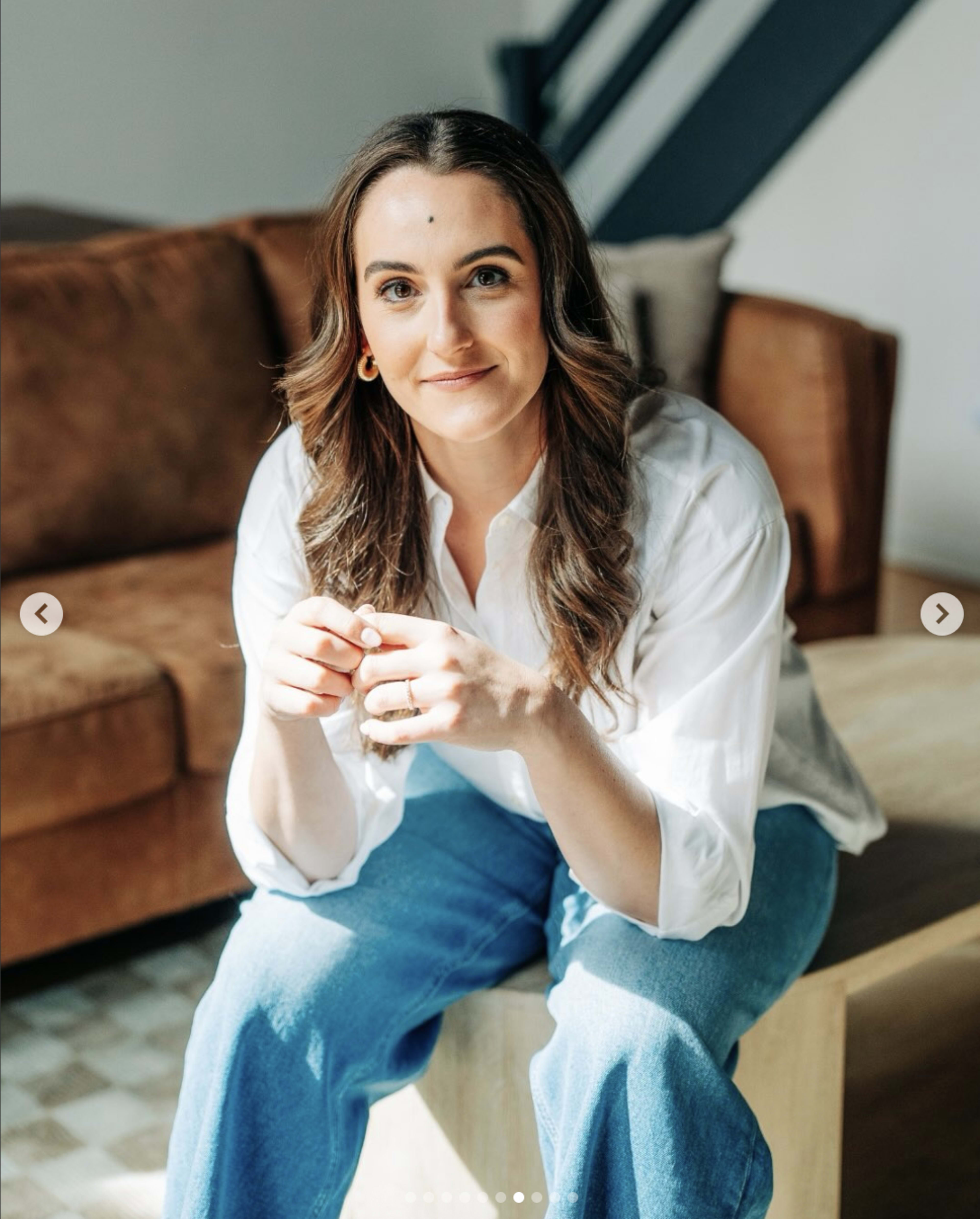 A woman with long hair is sitting on a coffee table in front of a brown sofa, wearing a white shirt and blue jeans, with a relaxed expression.