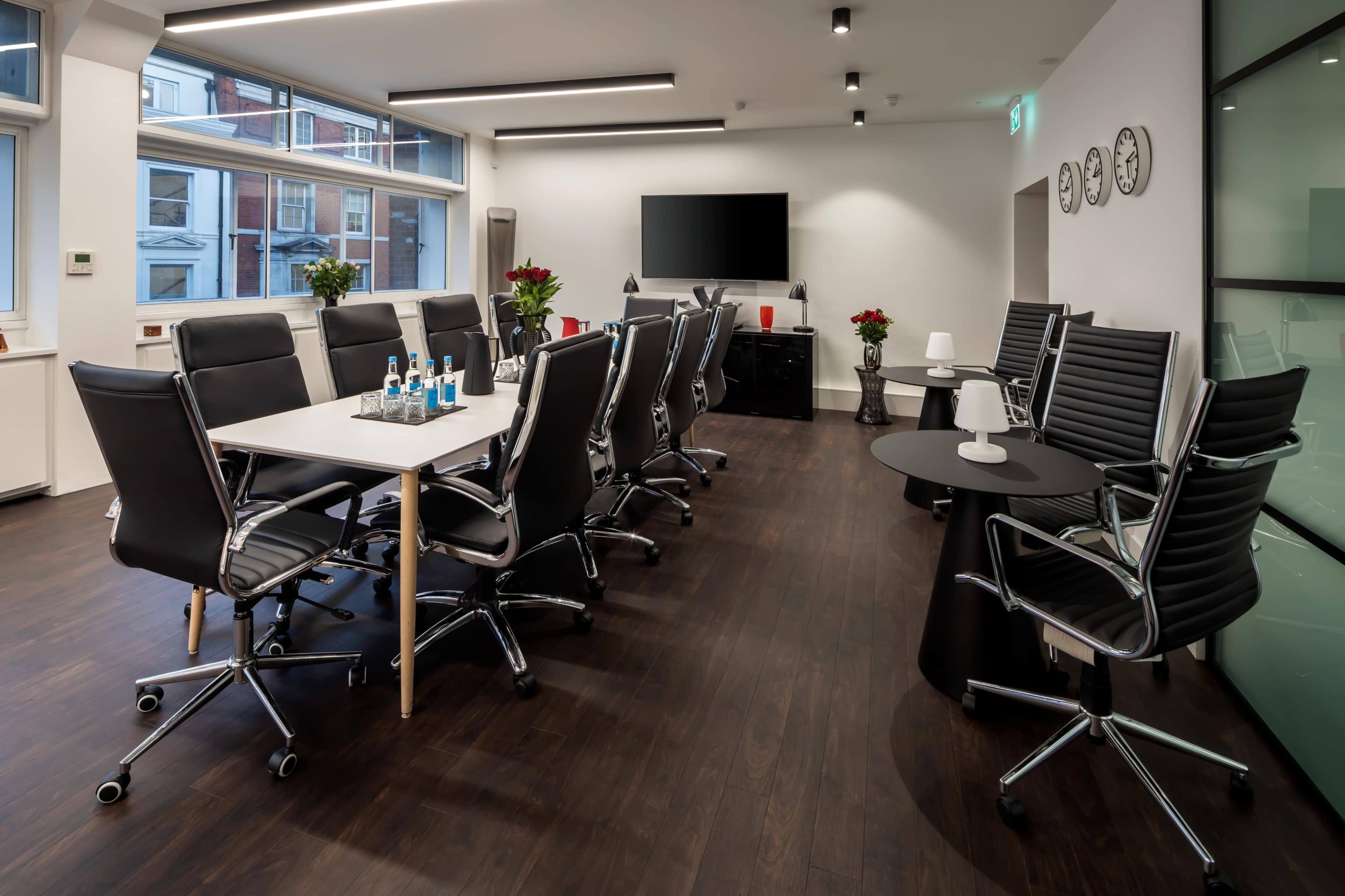 A modern conference room features a long table surrounded by ergonomic black chairs, with a television on the wall and decorative plants.