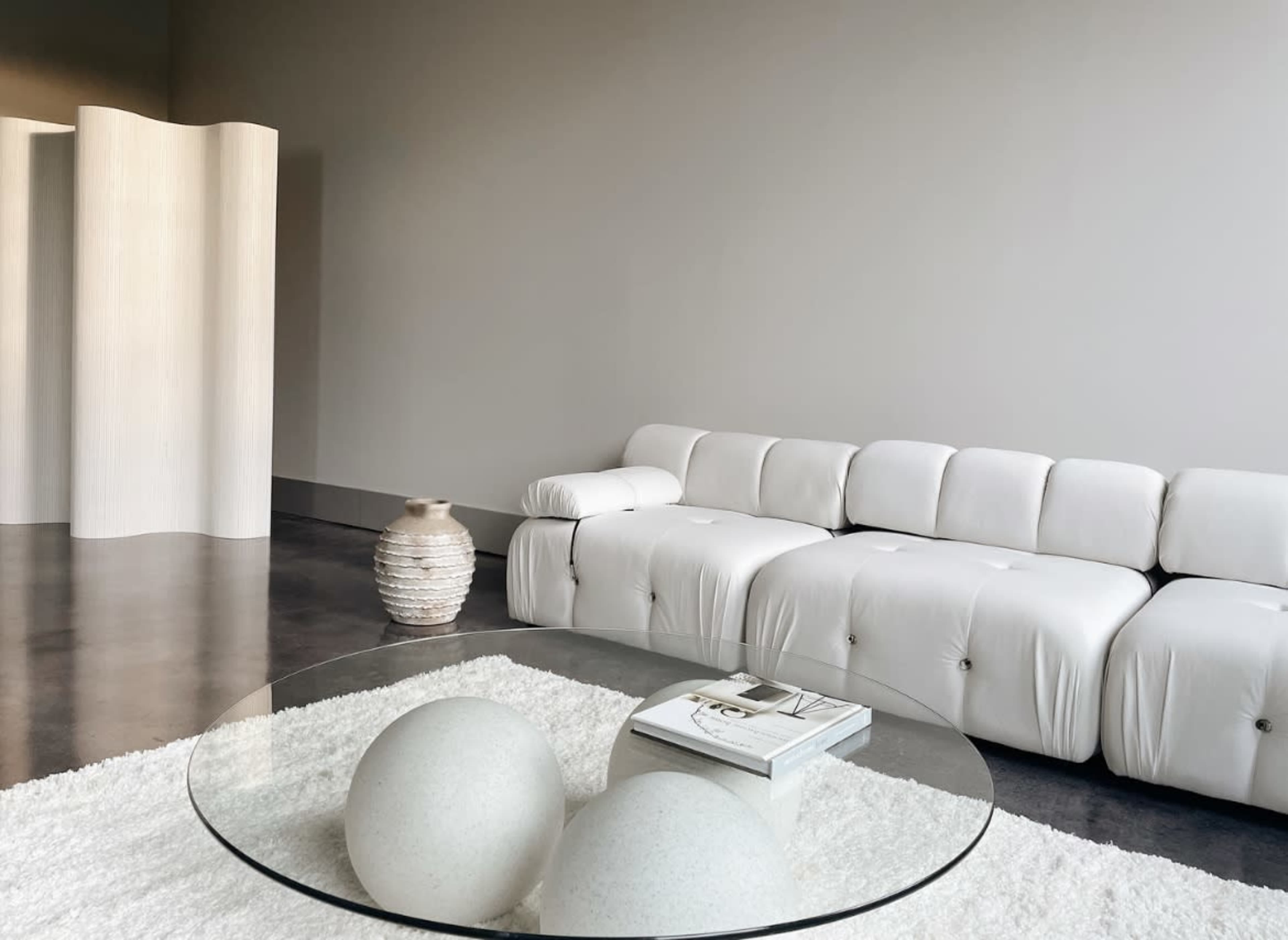 A modern, minimalist living space features a white sectional sofa, a circular glass coffee table with two spherical decor pieces, and a textured vase against a neutral wall.