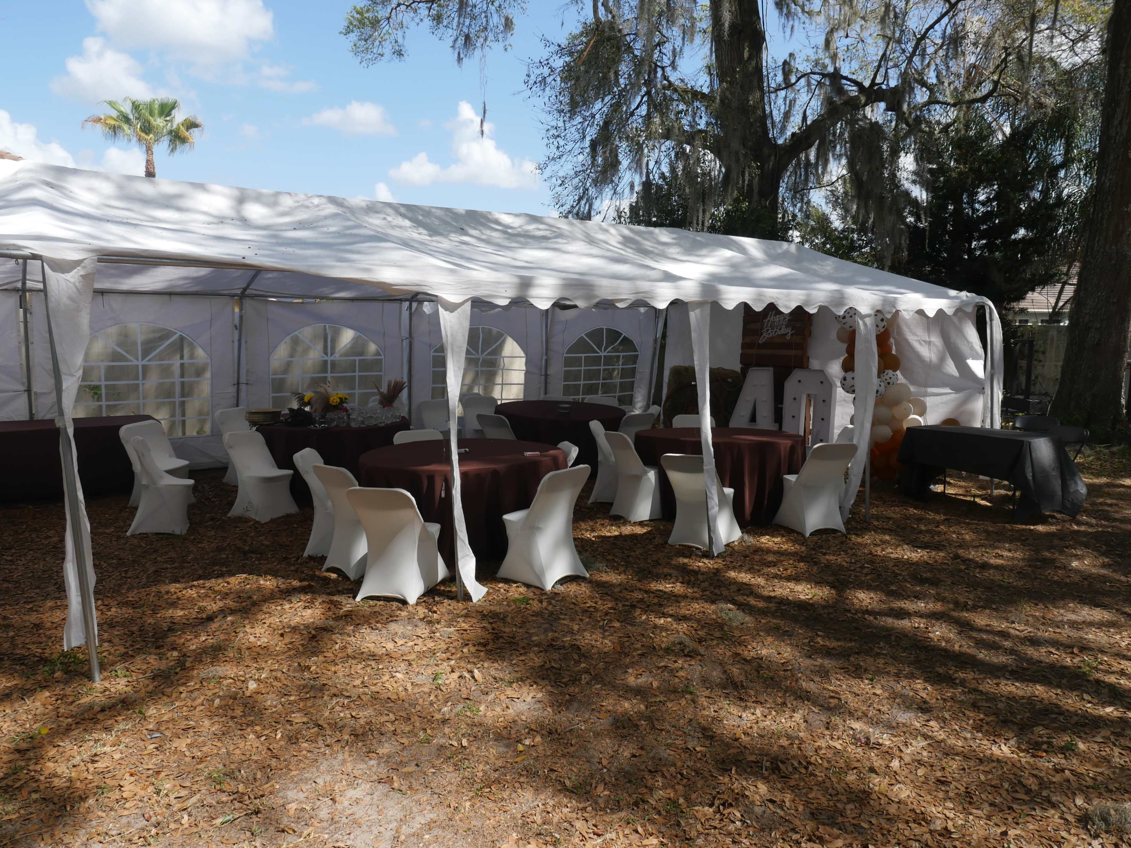 A tent with chairs and tables is set up in a shaded outdoor area covered with fallen leaves.