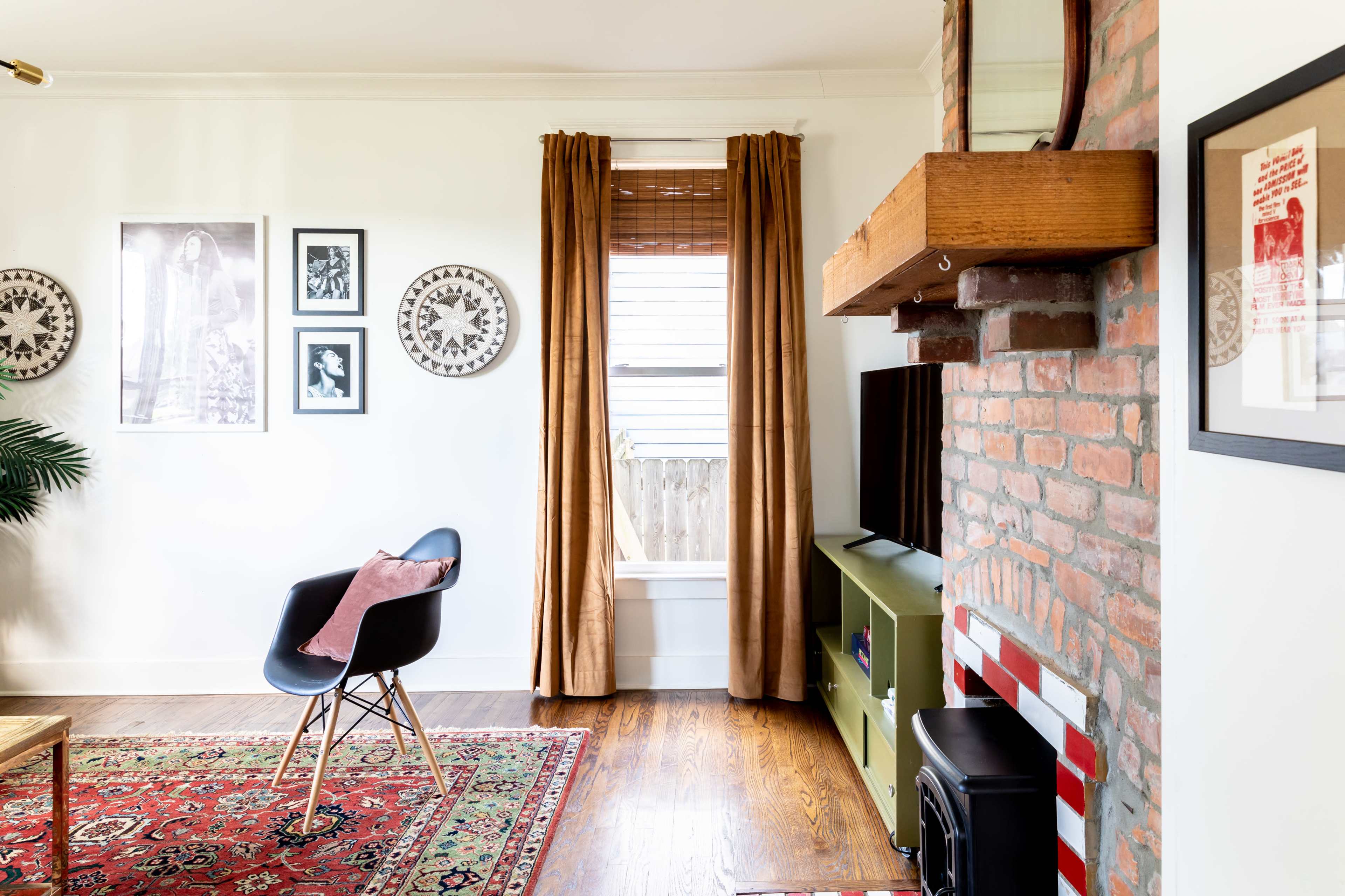 A living room features a brick fireplace, a television on an entertainment unit, framed artwork on the walls, and a window with brown curtains.