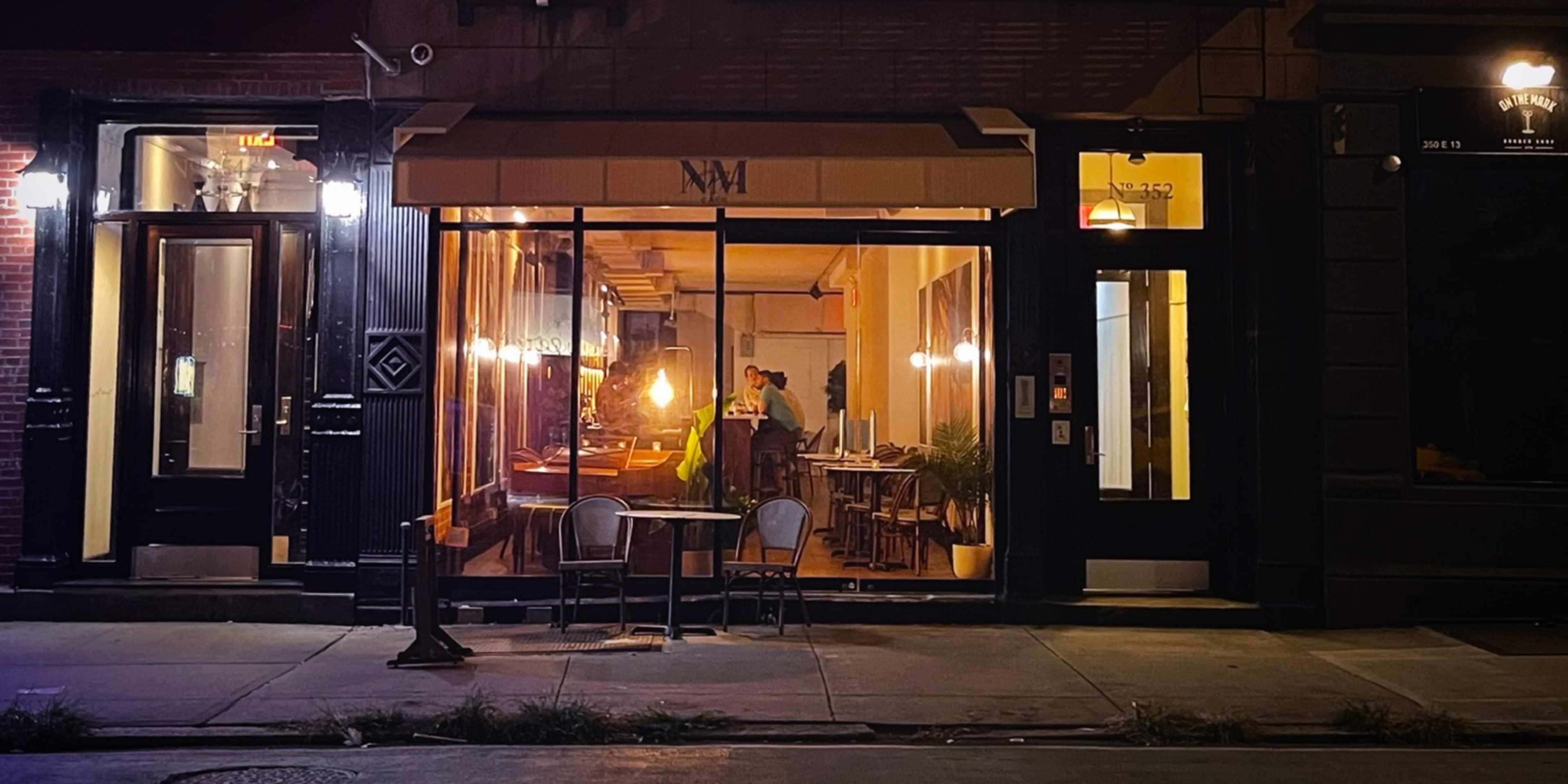 A restaurant with large front windows is illuminated at night, showing two people inside, with outdoor seating visible.