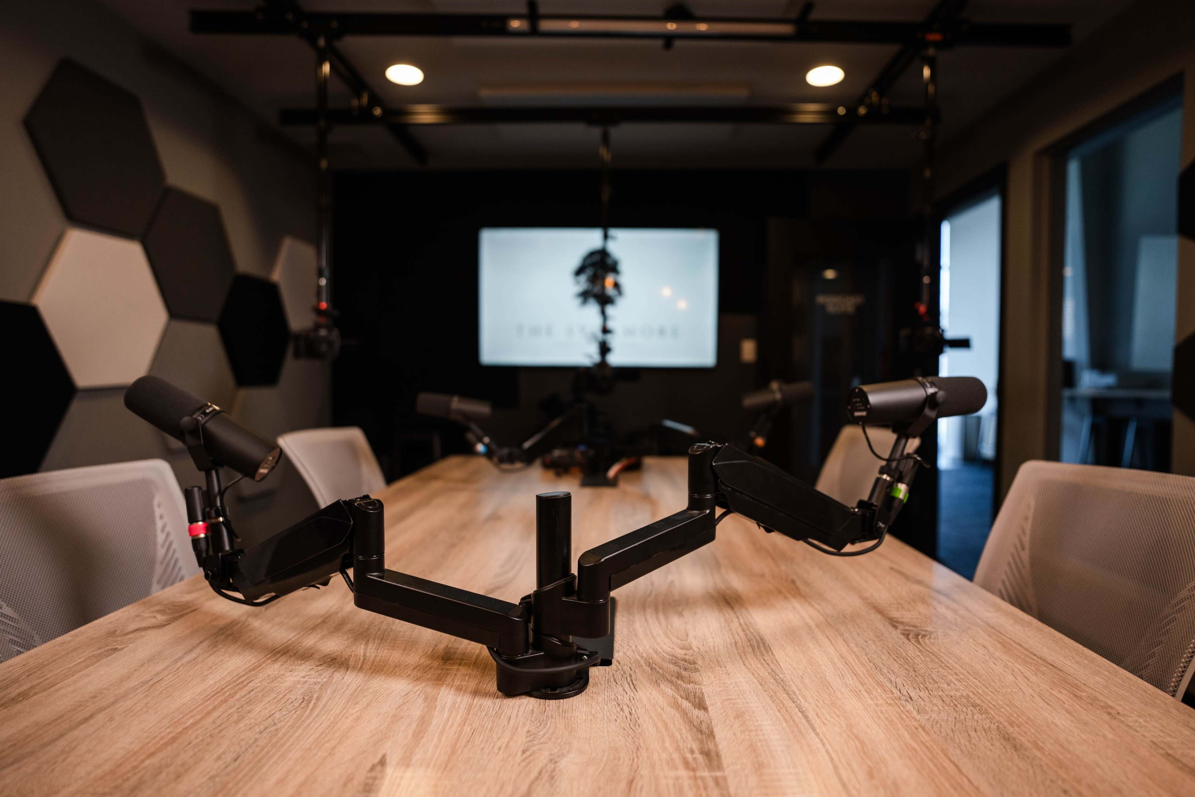 A modern conference room setup with a wooden table featuring multiple microphone arms positioned in front of a screen displaying a logo.