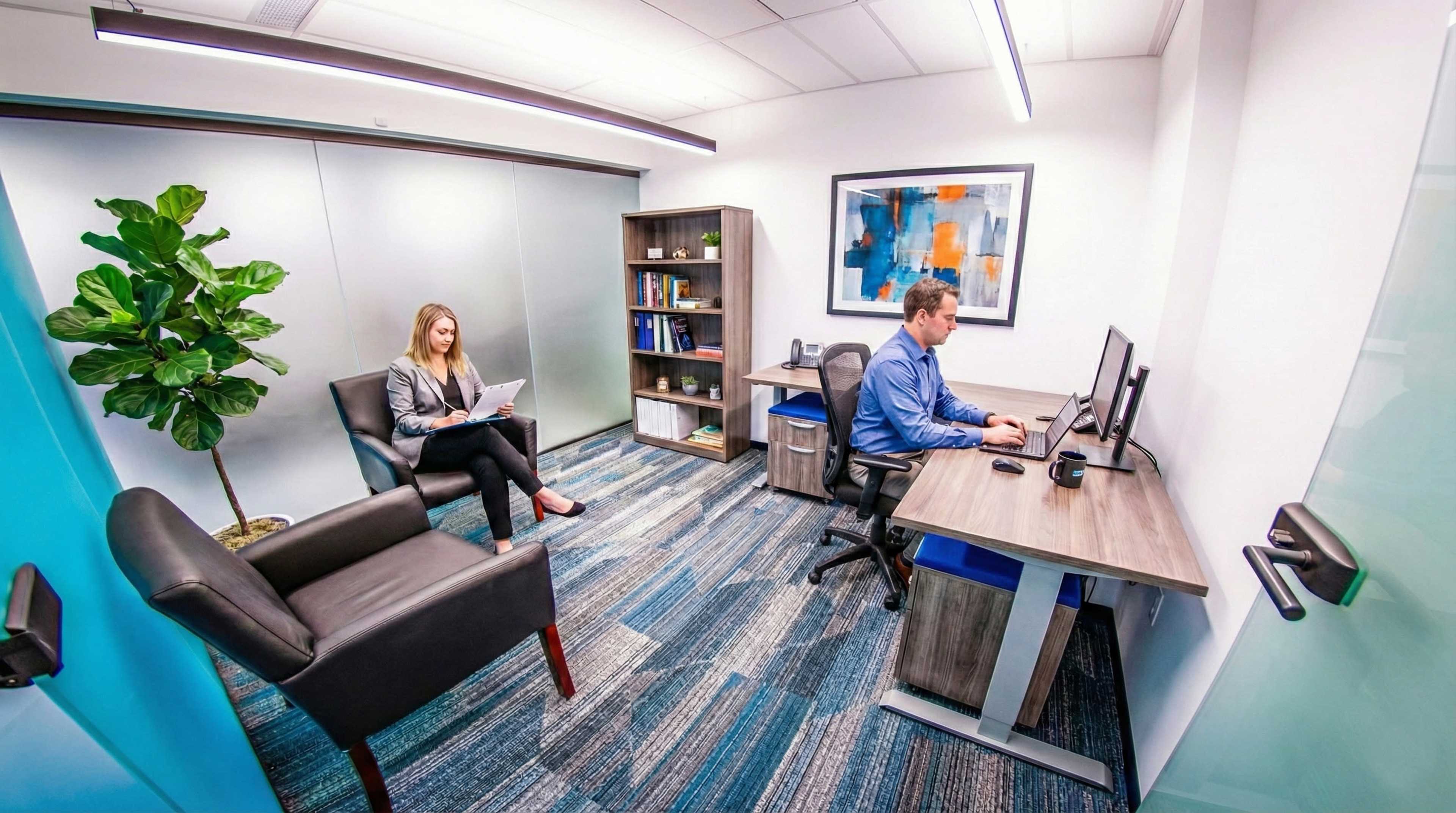 A modern office space features two individuals seated in separate areas, one working on a computer and the other using a laptop, with bookshelves and a large plant visible in the background.