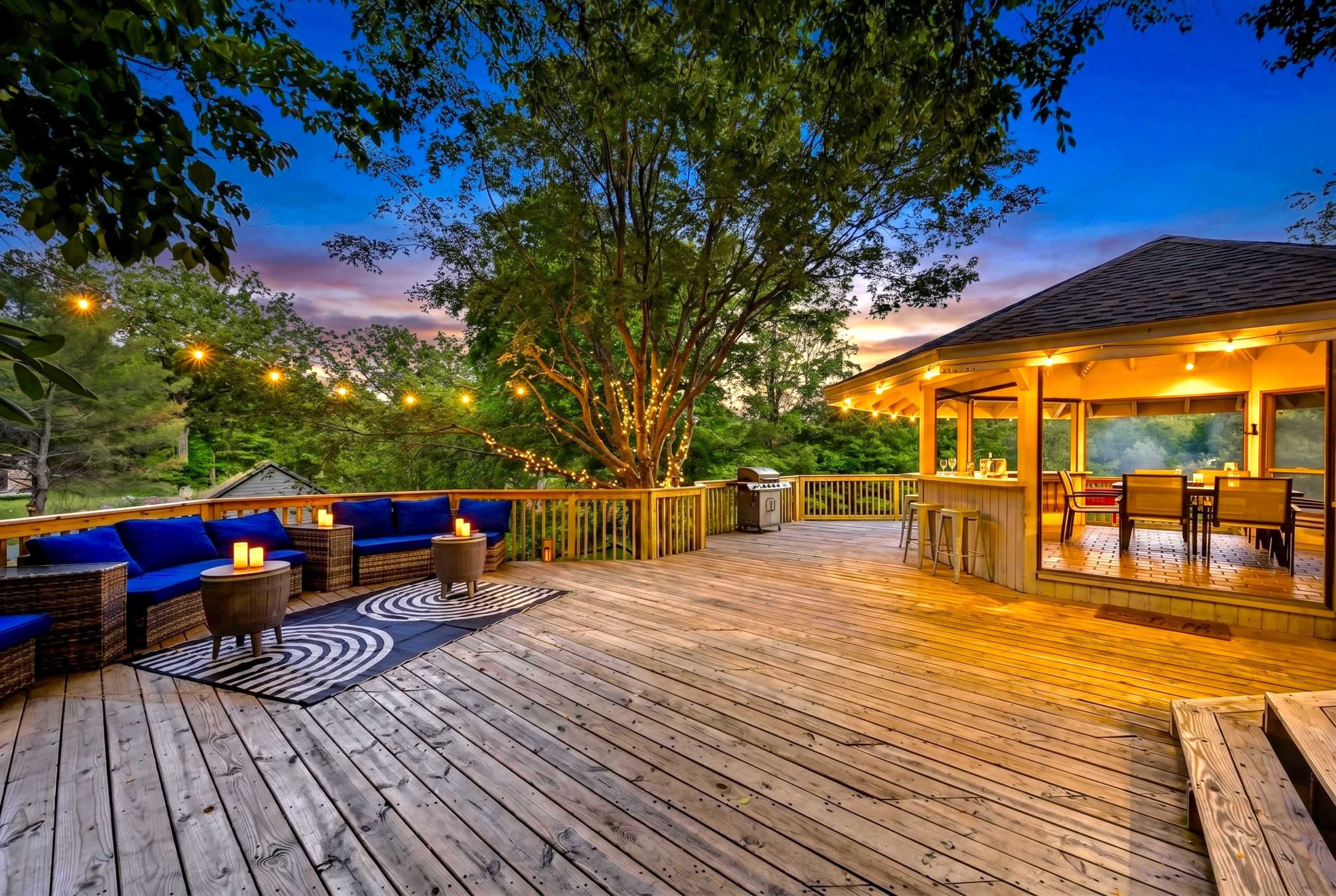 A spacious wooden deck features comfortable seating, a dining area under a gazebo, and string lights, surrounded by trees and a colorful sunset sky.