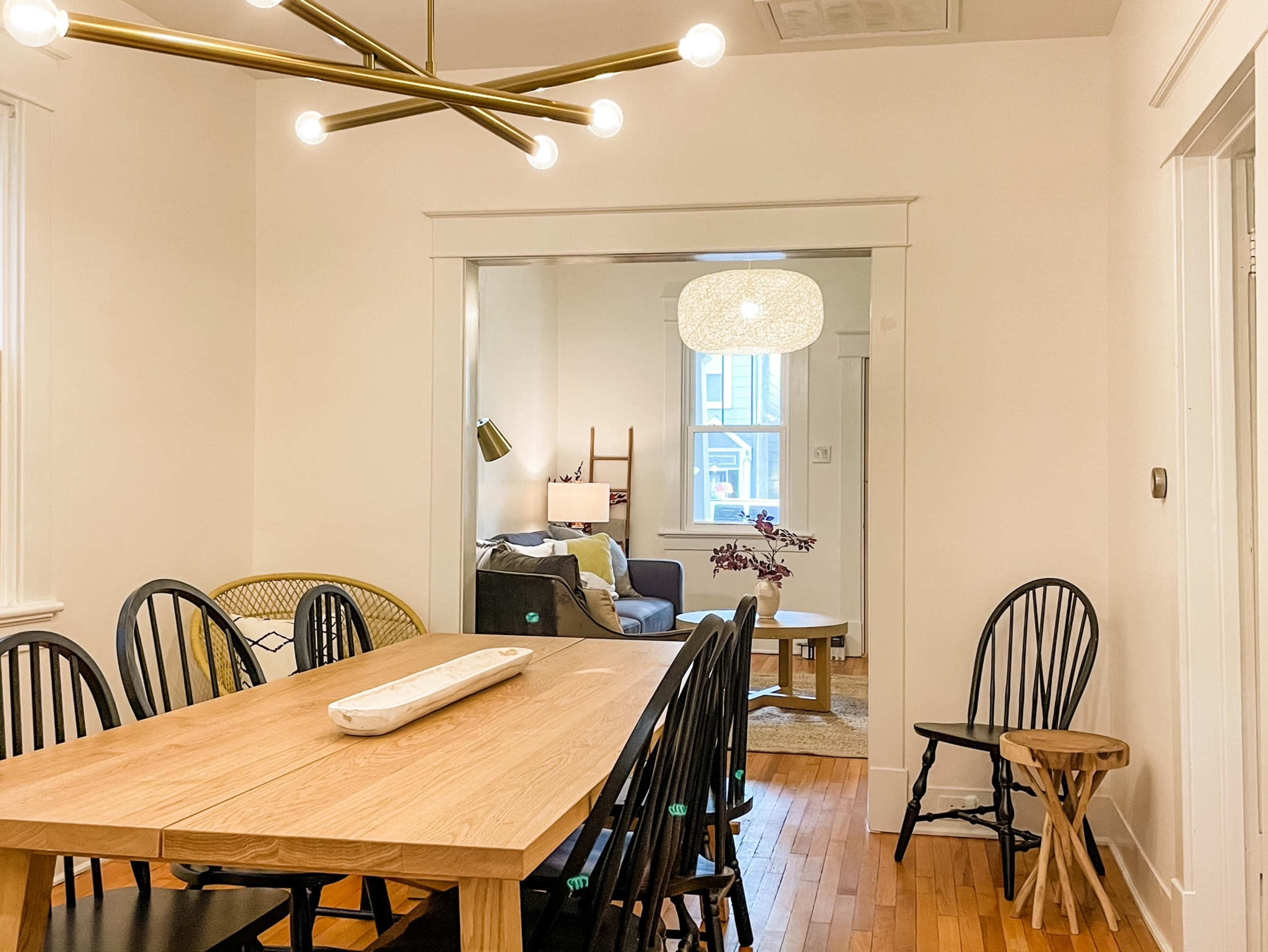 A dining room features a wooden table surrounded by black chairs, with a light fixture overhead and a view into a living area beyond.