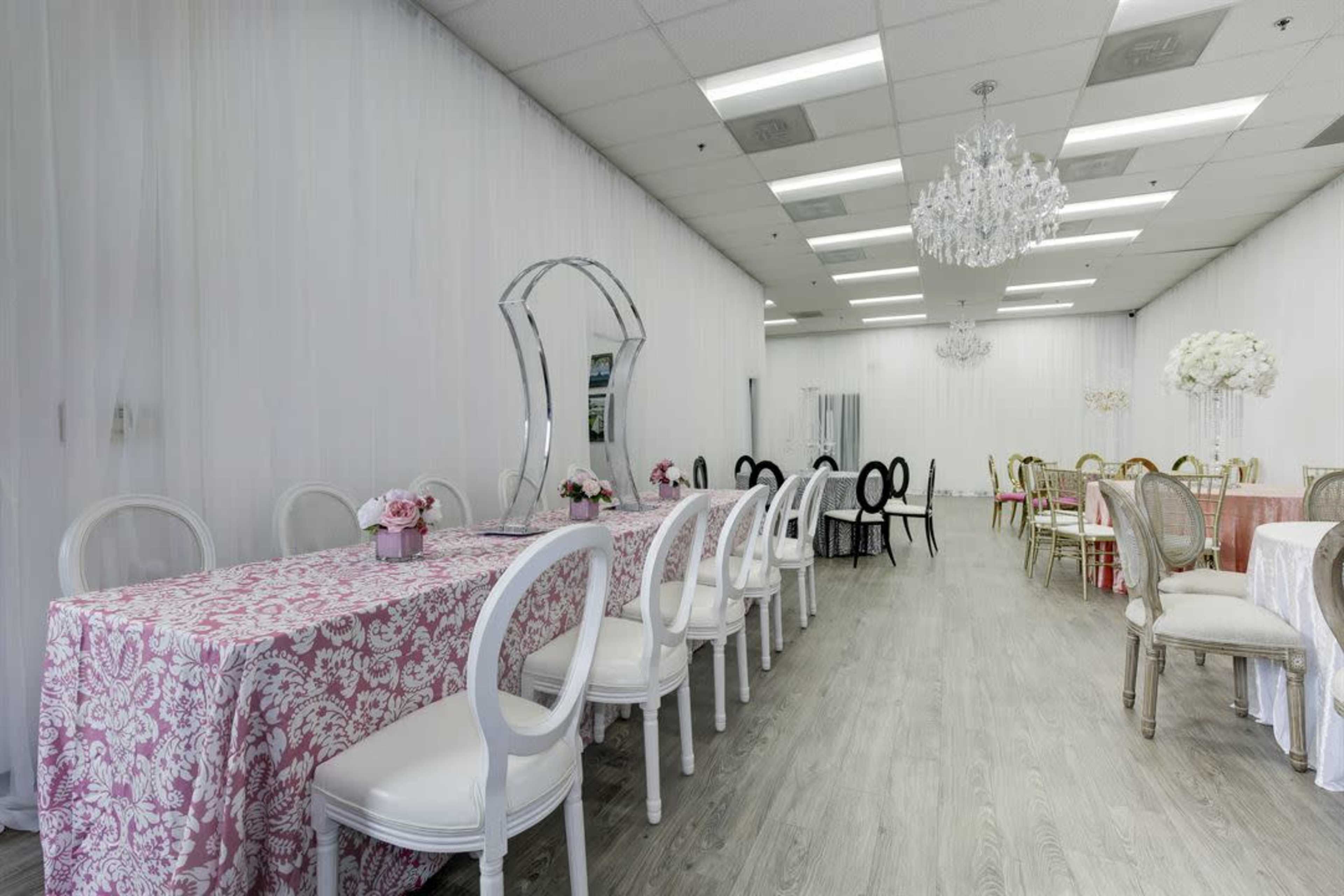The image shows a venue interior with several decorated tables and chairs arranged in a spacious area, featuring a chandelier and floral centerpieces.