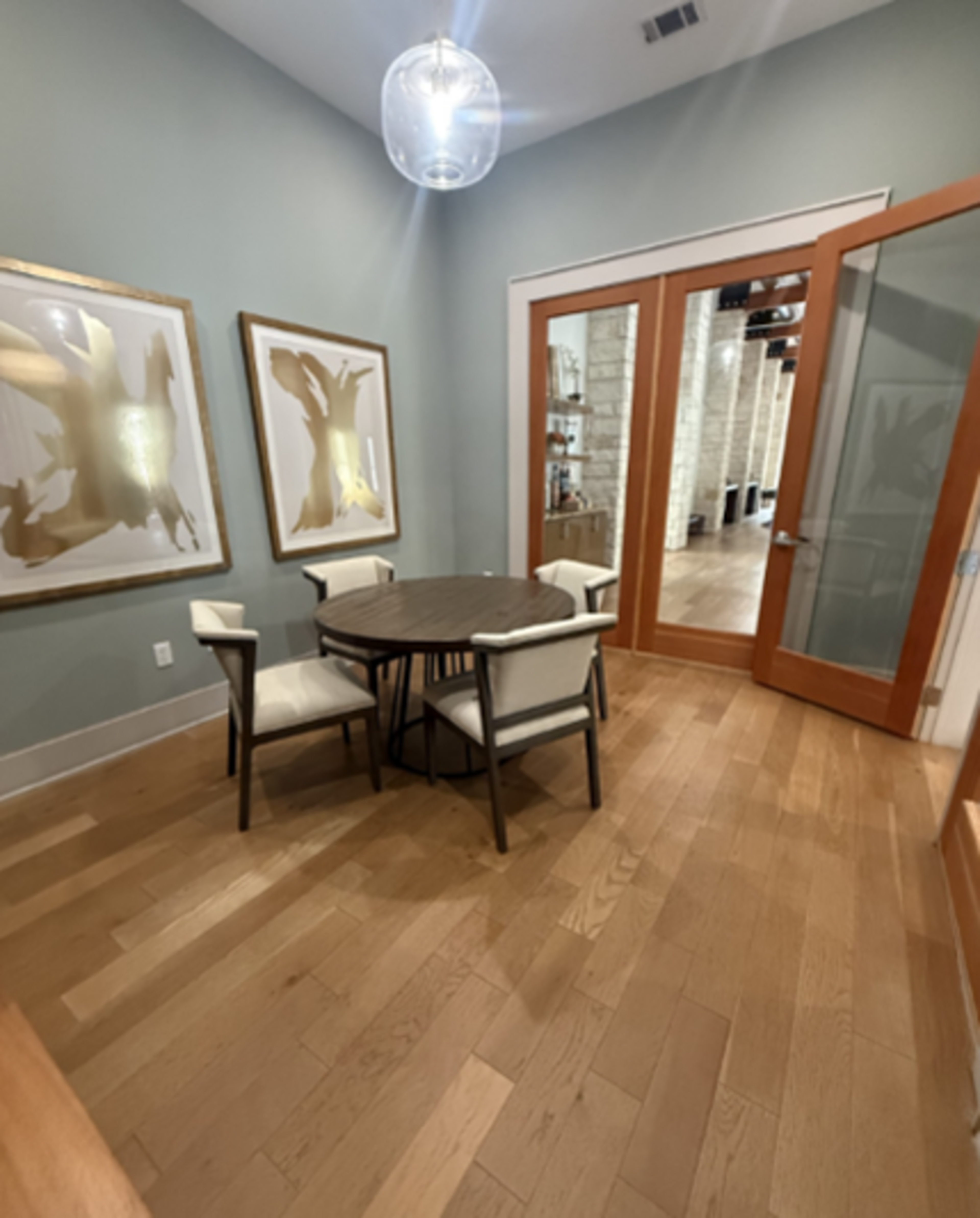 The image shows a small dining area with a circular table and four chairs, framed artwork on the walls, and glass doors leading to another room.