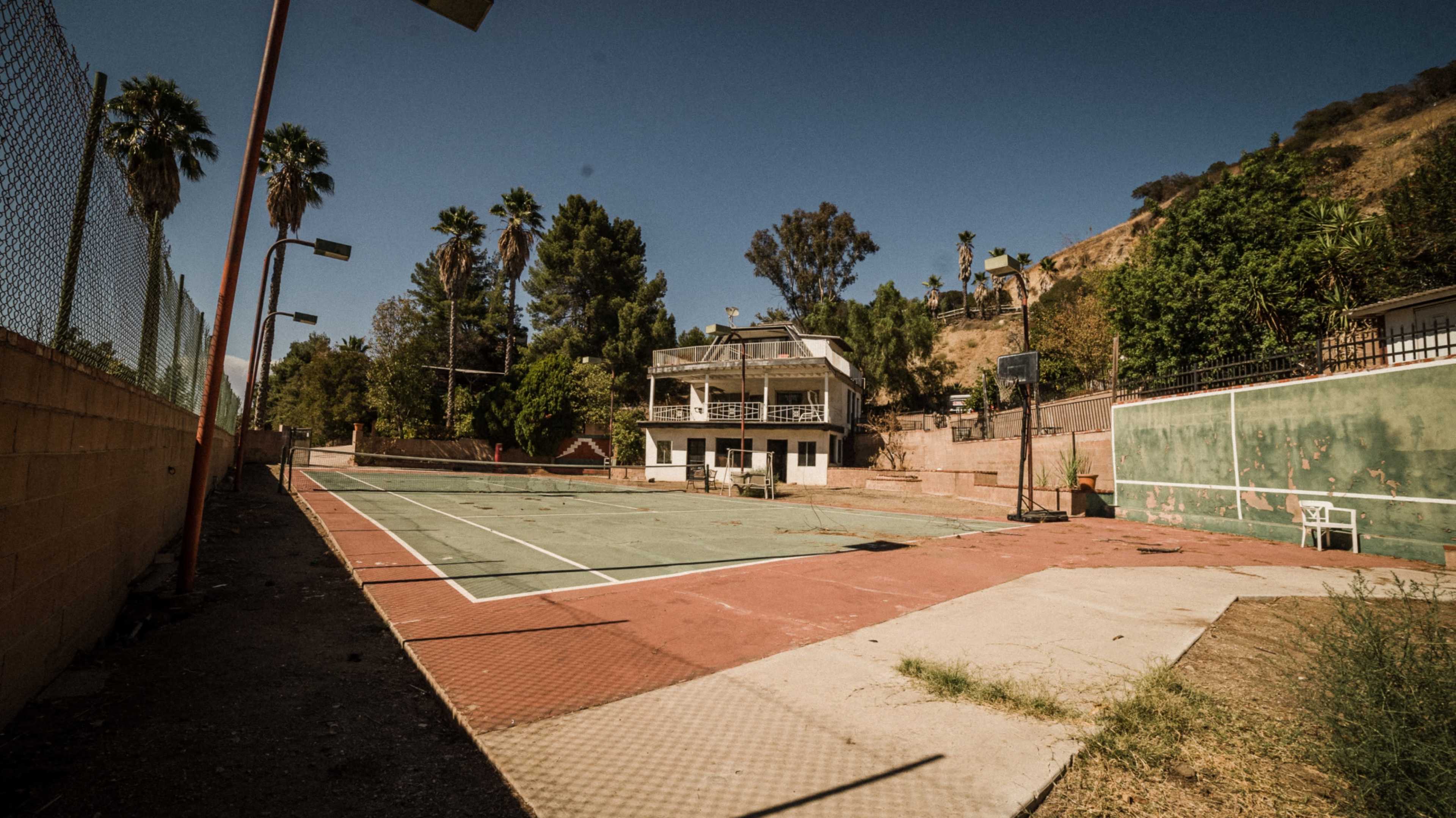 An abandoned outdoor basketball court sits beside a derelict two-story building, surrounded by palm trees and a hillside.