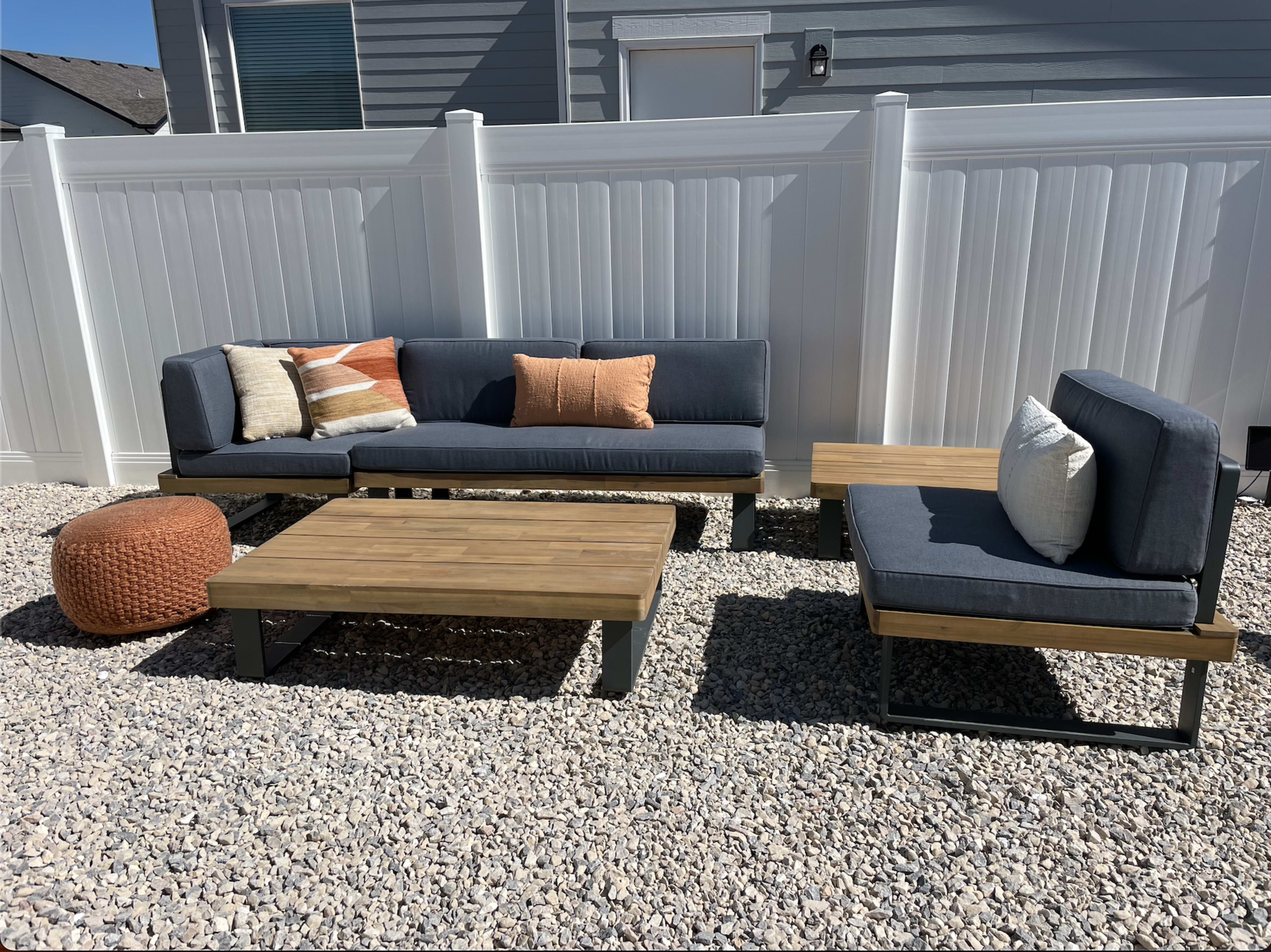 A modern outdoor seating area features a sectional sofa, a single chair, and a coffee table made of wood, all placed on gravel beneath a white vinyl fence.