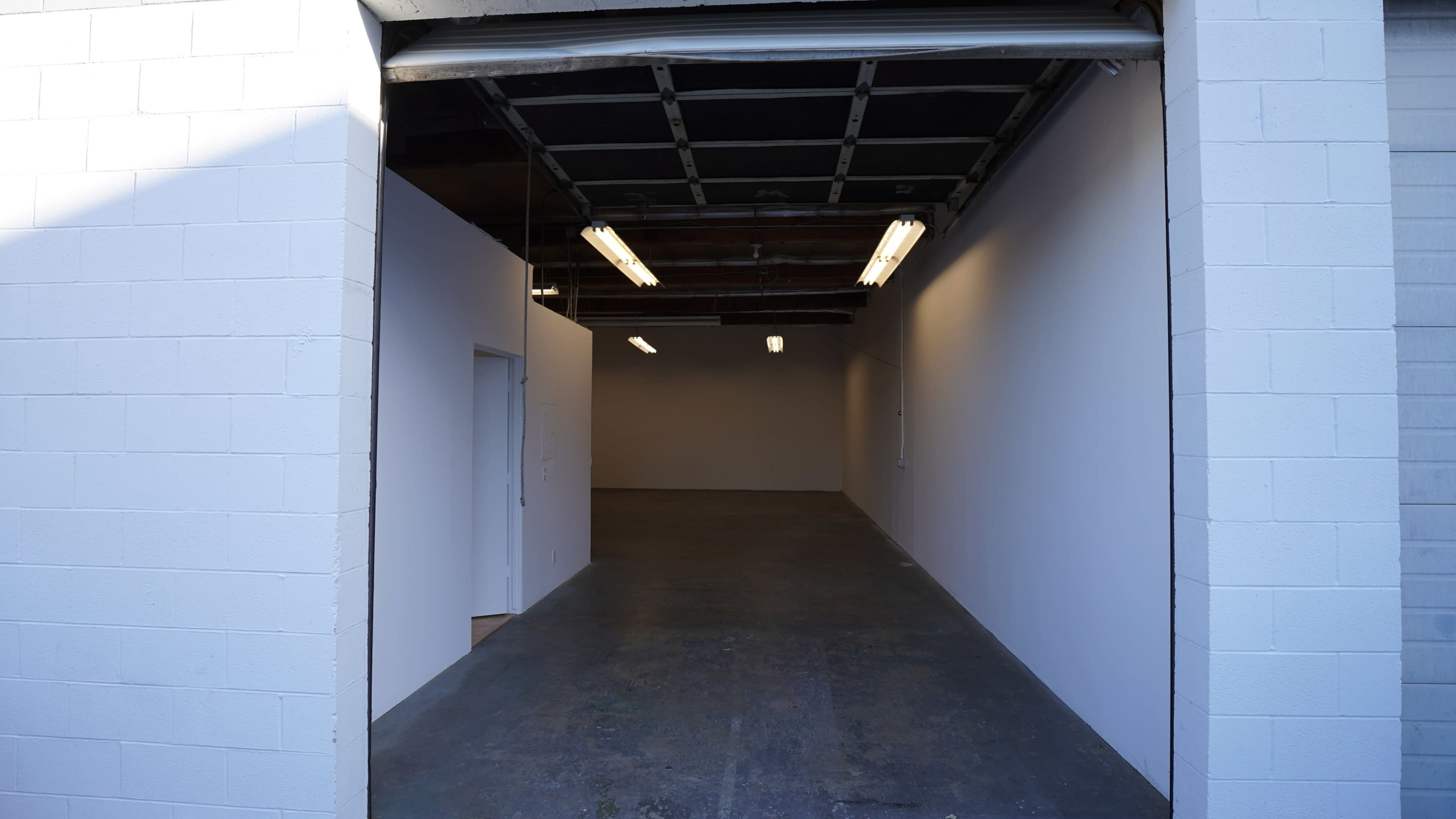 The image shows a large, empty room with white walls and concrete flooring viewed through an open garage door.