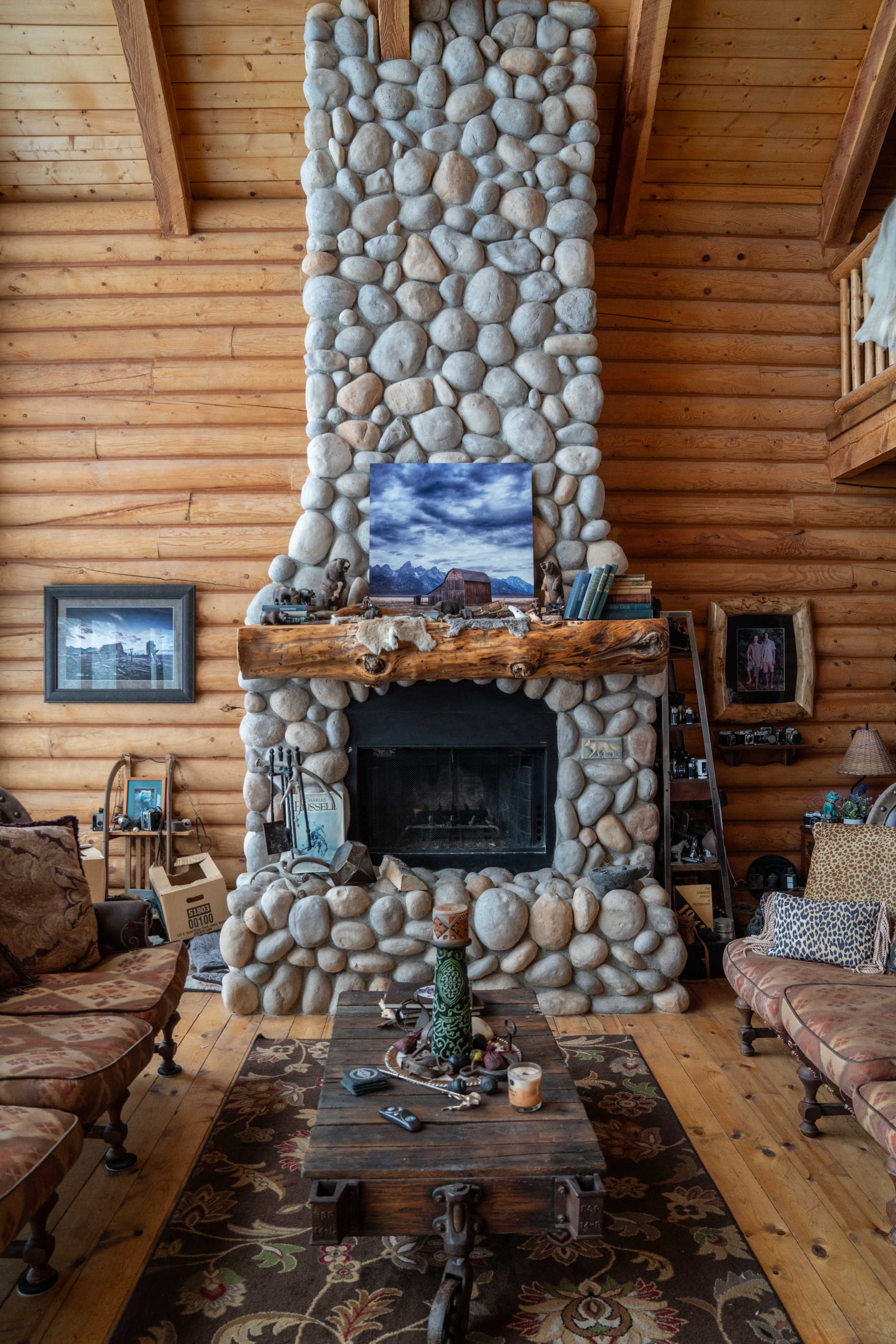 A rustic living room featuring a stone fireplace, a wooden mantel with decorations, and a wooden table surrounded by upholstered seating.